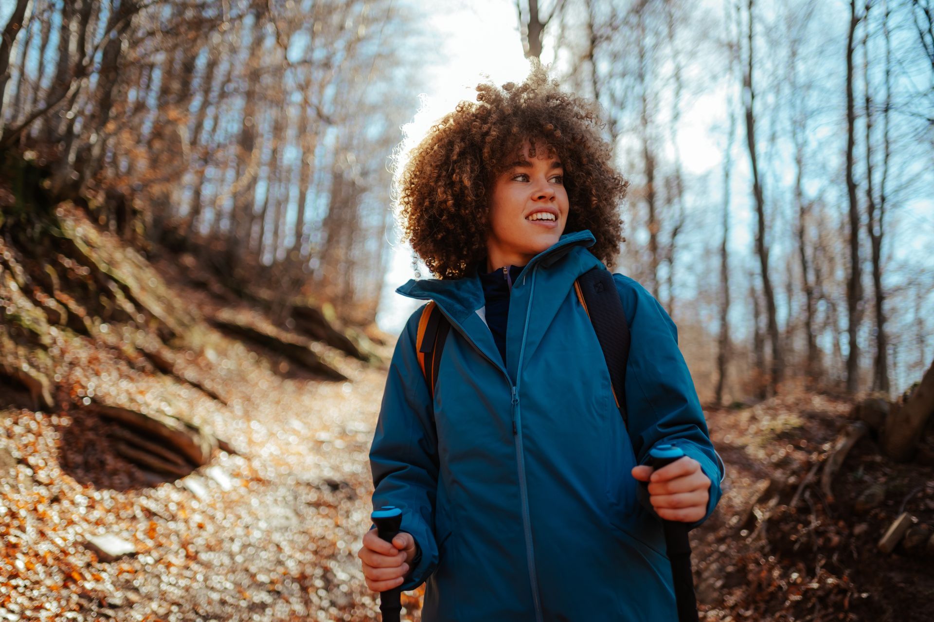 Woman hikes in forest, wearing blue jacket, holding poles, smiling, looking up.