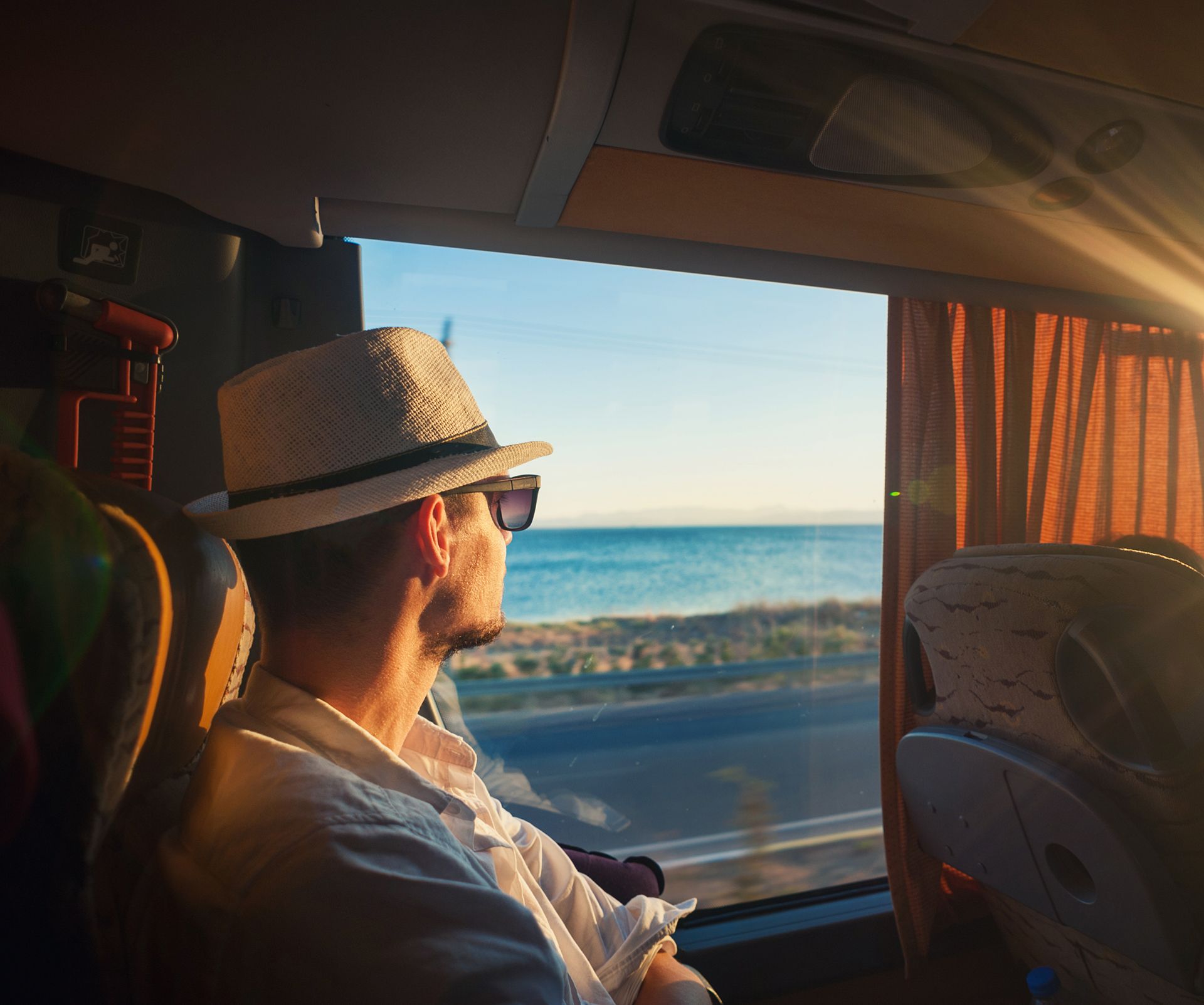 Man in hat and sunglasses looking out bus window at ocean.