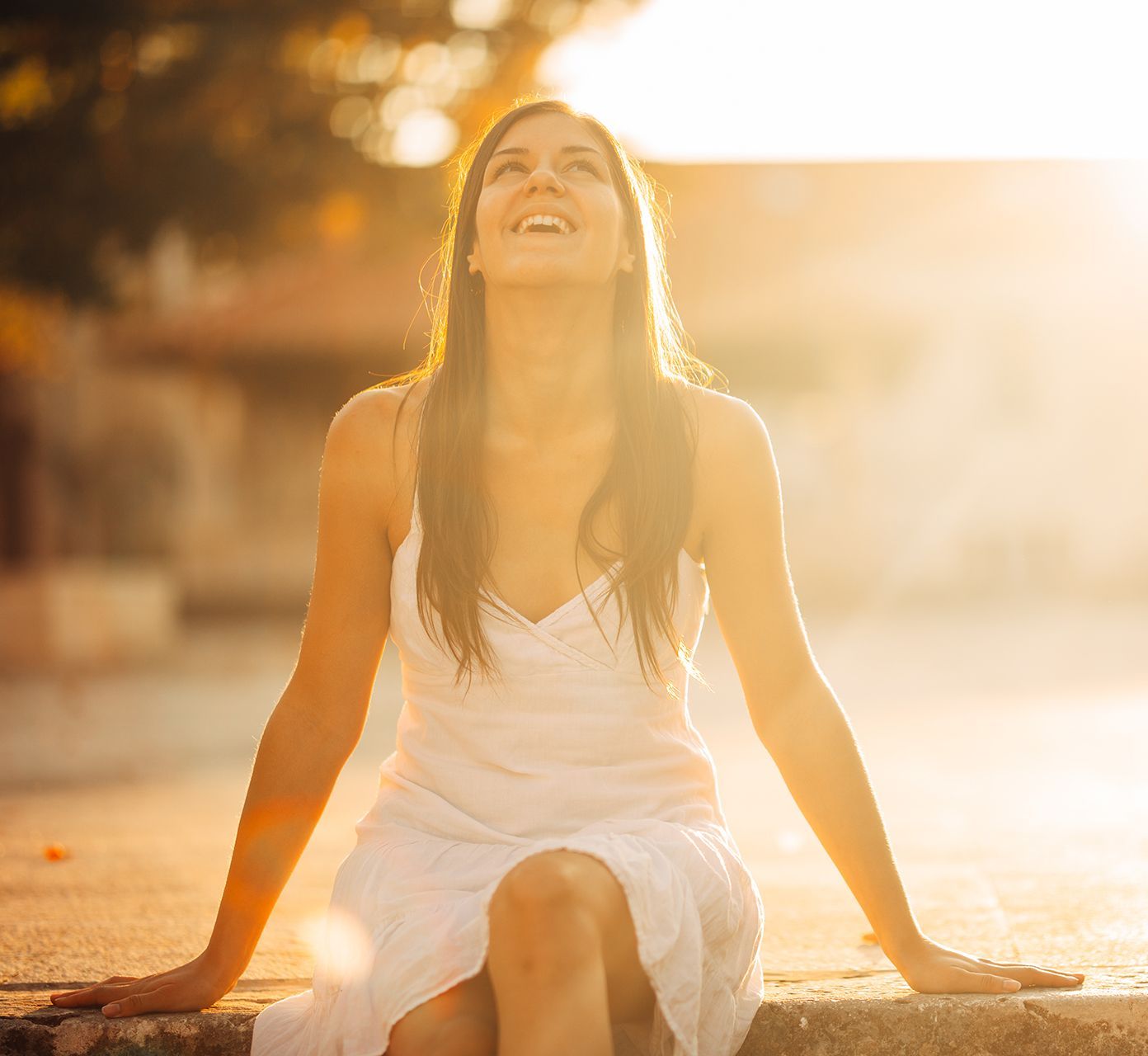 A woman in a white dress is sitting on a curb looking up at the sun