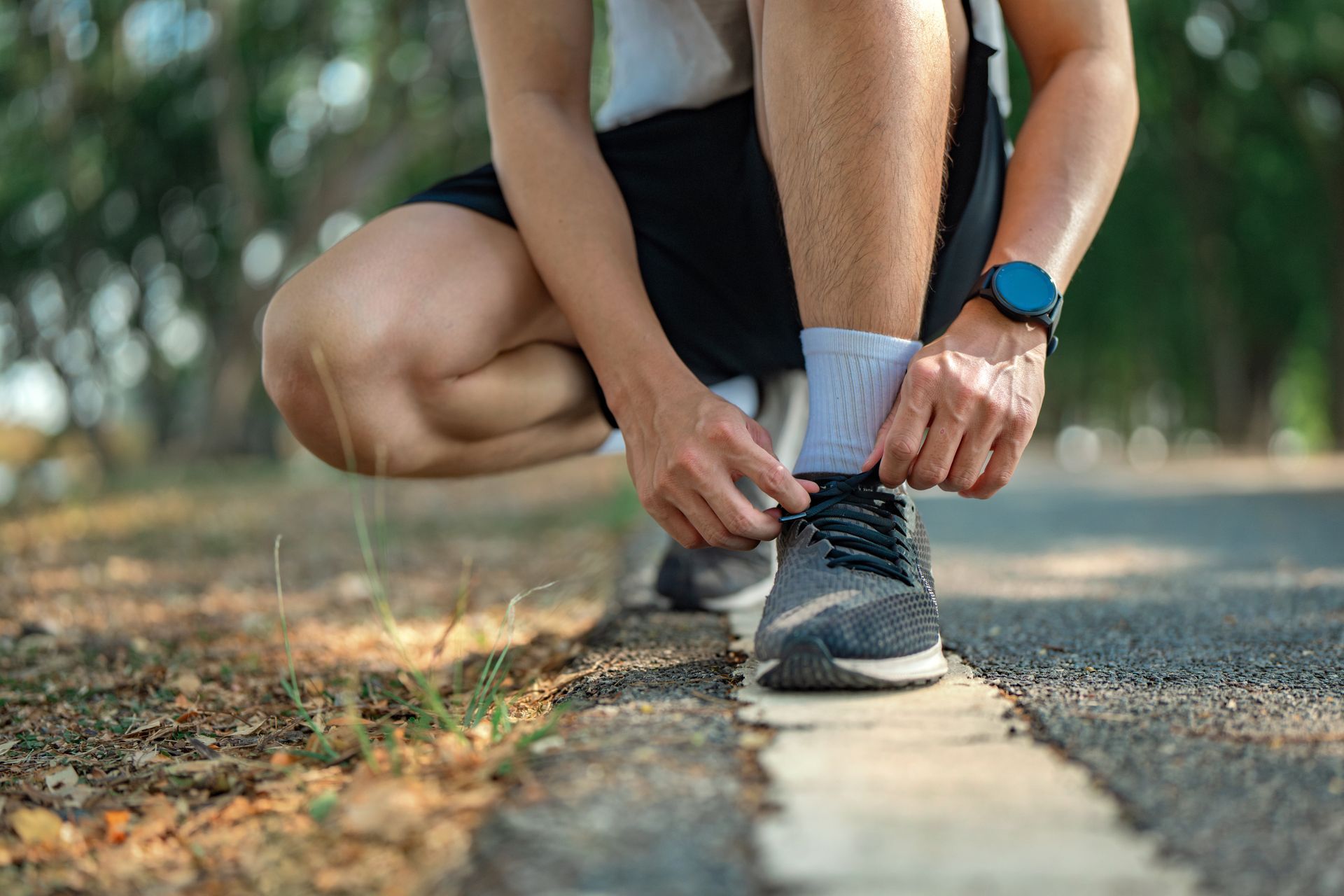 Person kneeling, tying running shoe on a paved path, wrist watch visible.