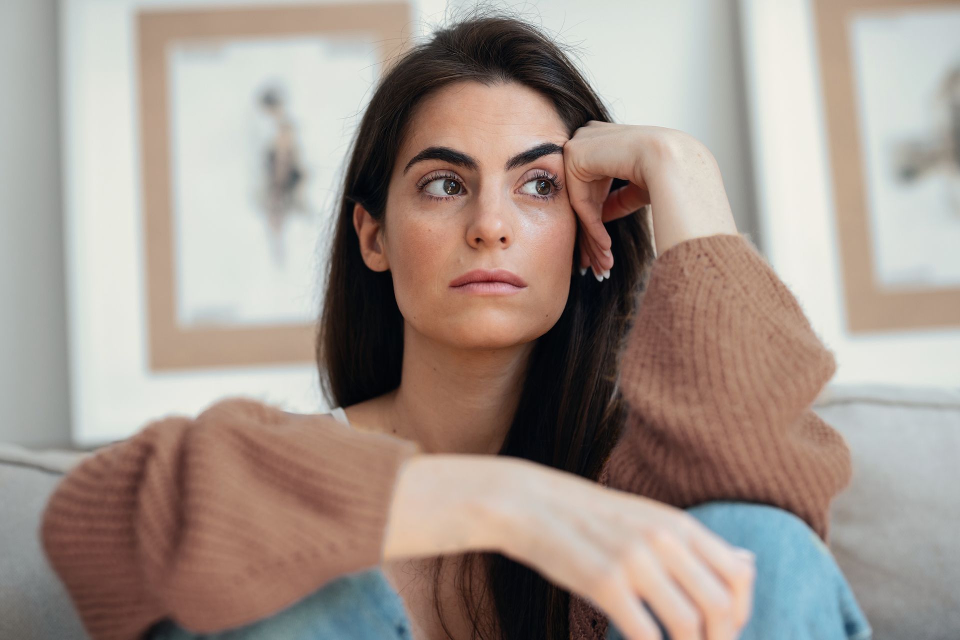 Woman with dark hair, looking thoughtful, resting head on hand. Brown sweater, sitting on a couch.