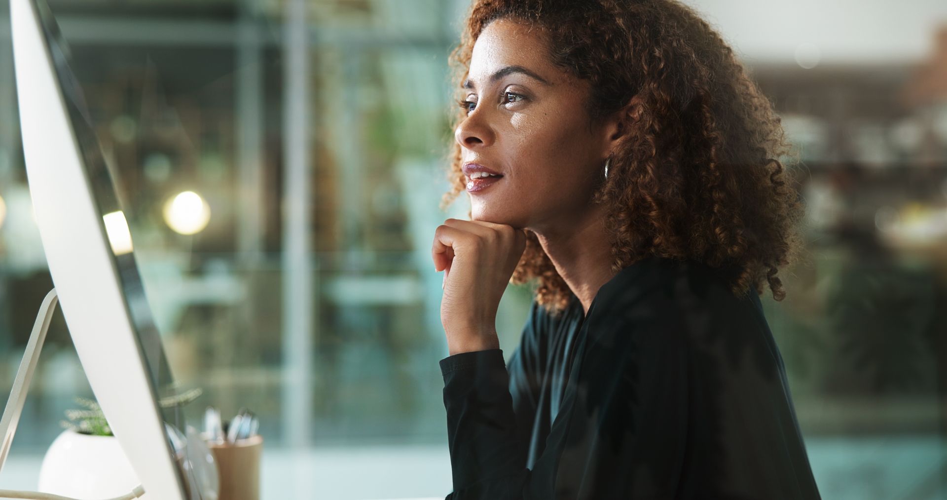 Woman thoughtfully looking at computer screen, hand on chin, in office setting.