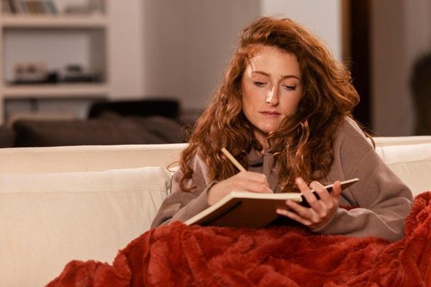 Woman with red hair writing in a notebook on a sofa, covered by a red blanket.
