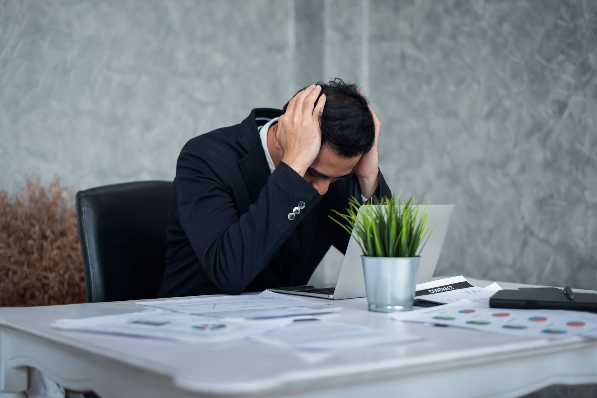 Man in suit, head in hands at desk, stressed by paperwork, next to a laptop and plant.