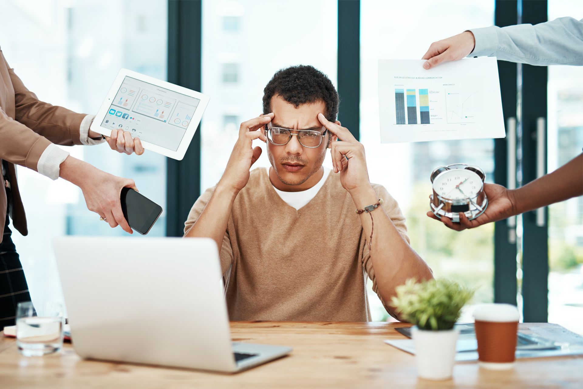 Man at desk overwhelmed, holding head; hands hold tablet, charts, phone, and clock.