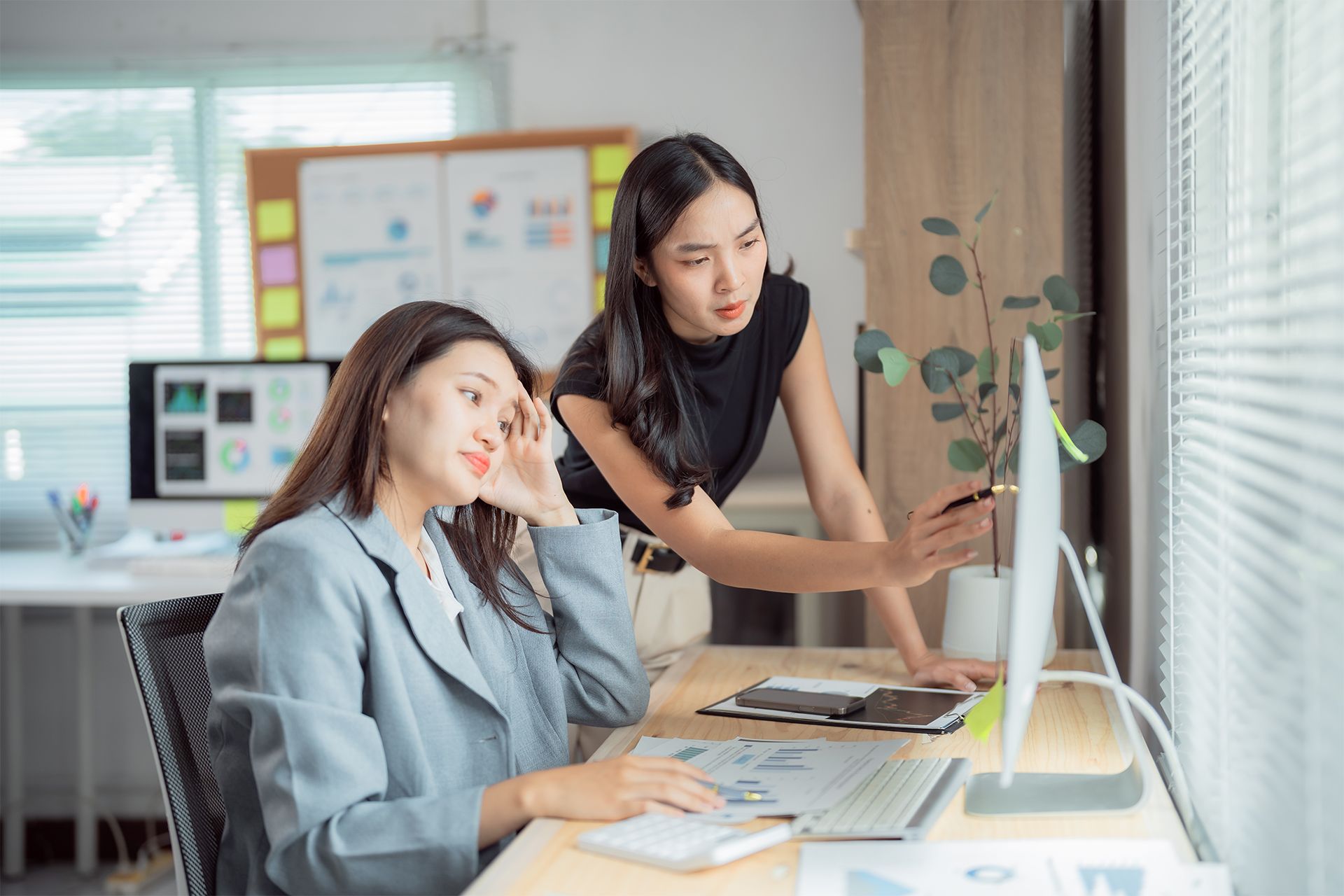 Two women collaborate at a desk, looking at a computer screen in an office.