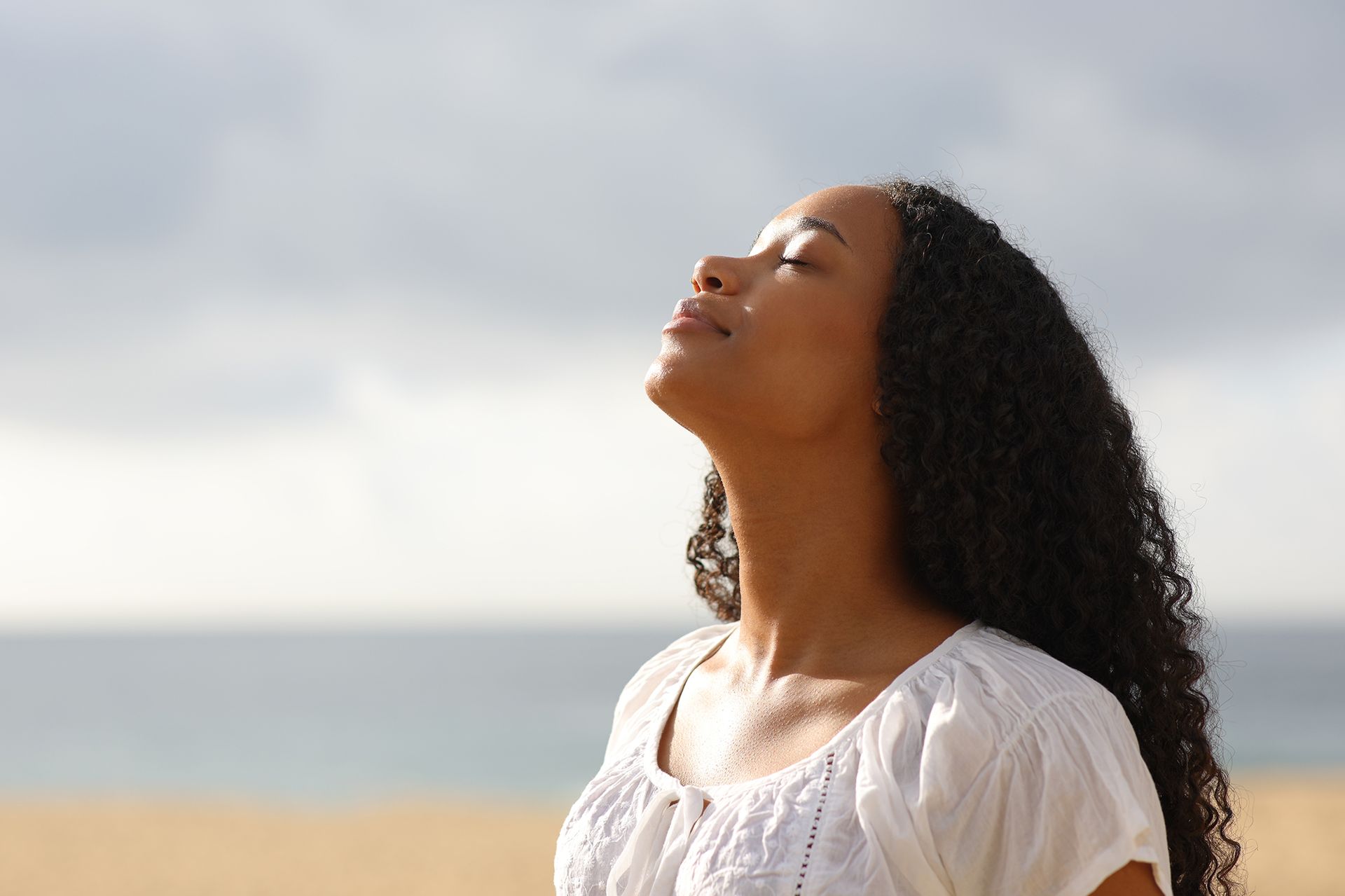 Woman with curly hair, eyes closed, tilts head back and inhales deeply on a beach, against a seascape.