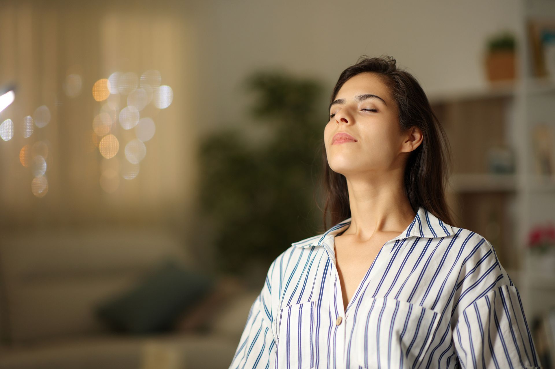 Woman in striped shirt with eyes closed, breathing deeply in a softly lit room.