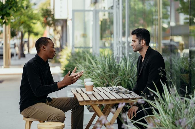 Two people talking at a small outdoor café table with coffee cups and greenery nearby.