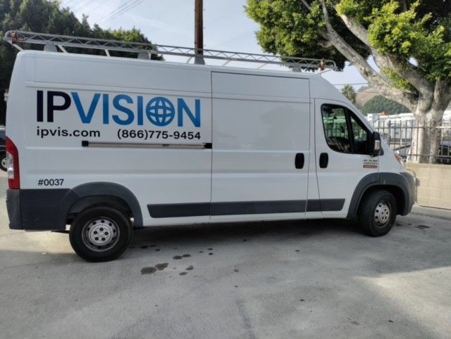 White IPVision van with blue logo, parked on a street. Ladder on the roof.