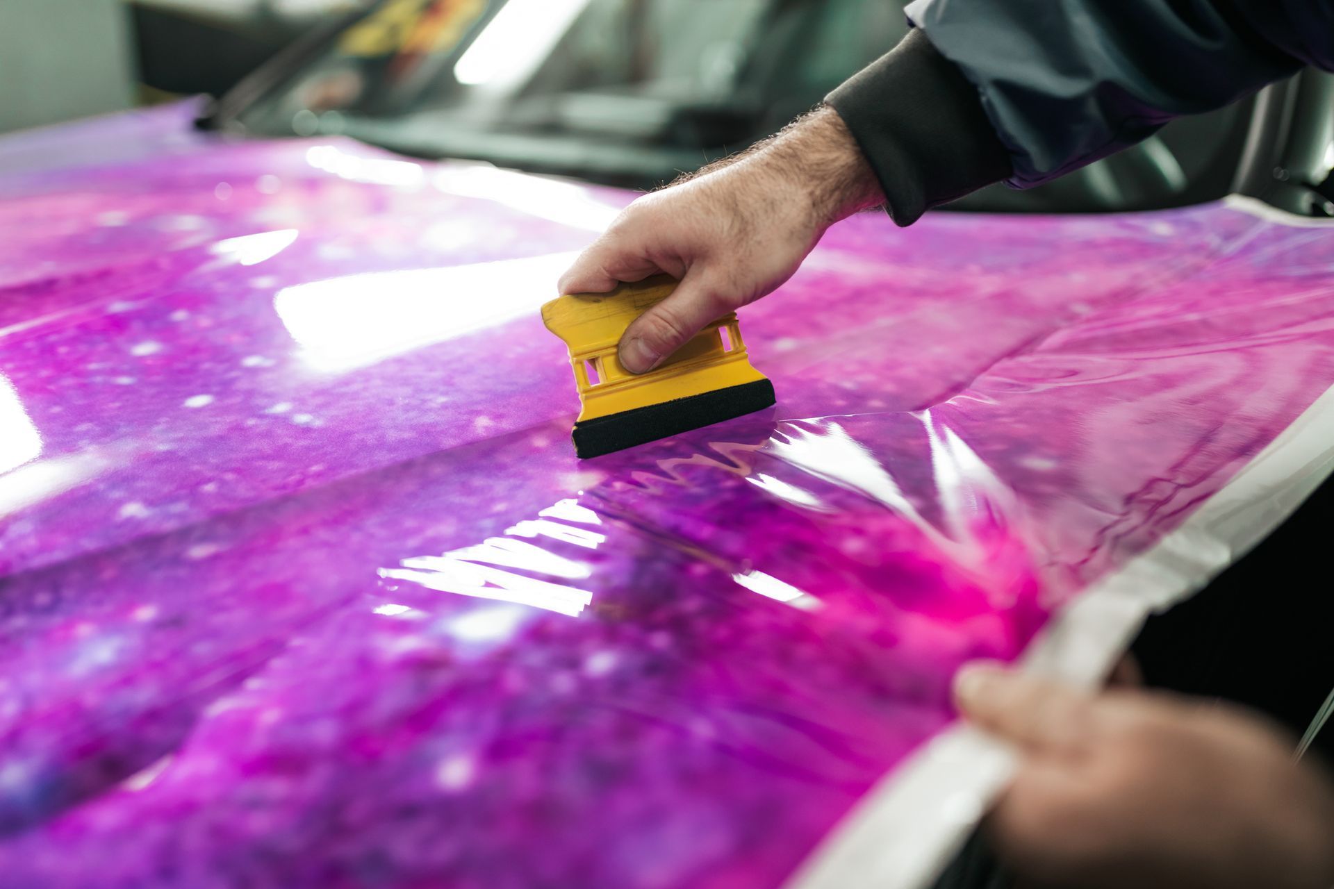 Person applying a galaxy-patterned wrap to a car hood, using a yellow squeegee.