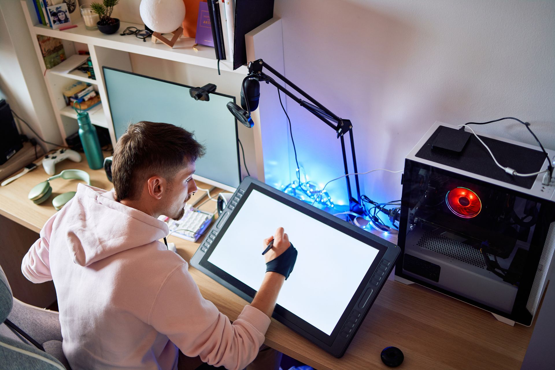 Man drawing on a digital tablet at a desk with two monitors, in a room with gaming gear.