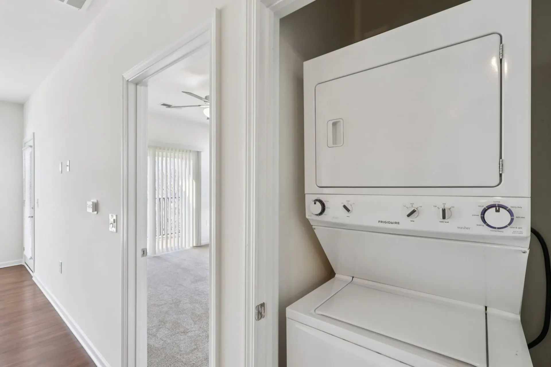 Stackable washer and dryer in a hallway with a bedroom visible through an open door.