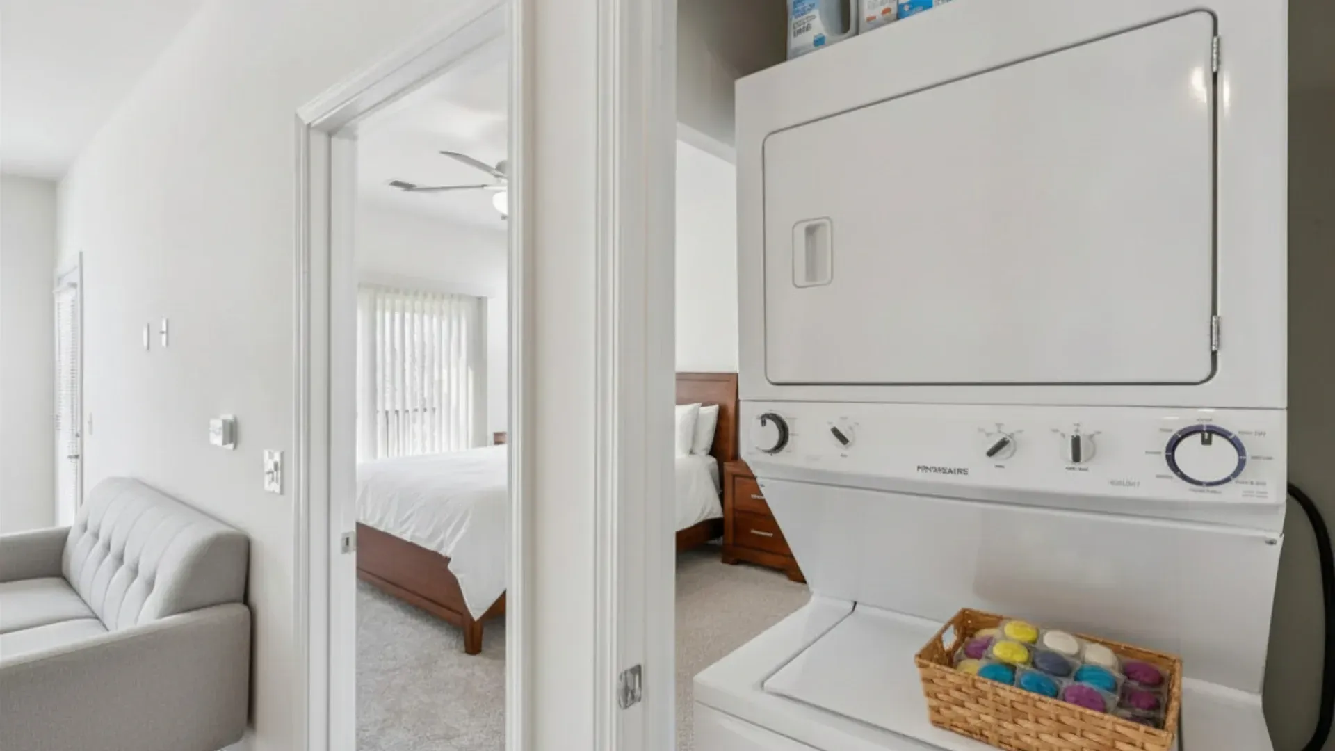 Stacked washer and dryer with a basket of laundry pods, open doorway to bedroom with bed and ceiling fan.