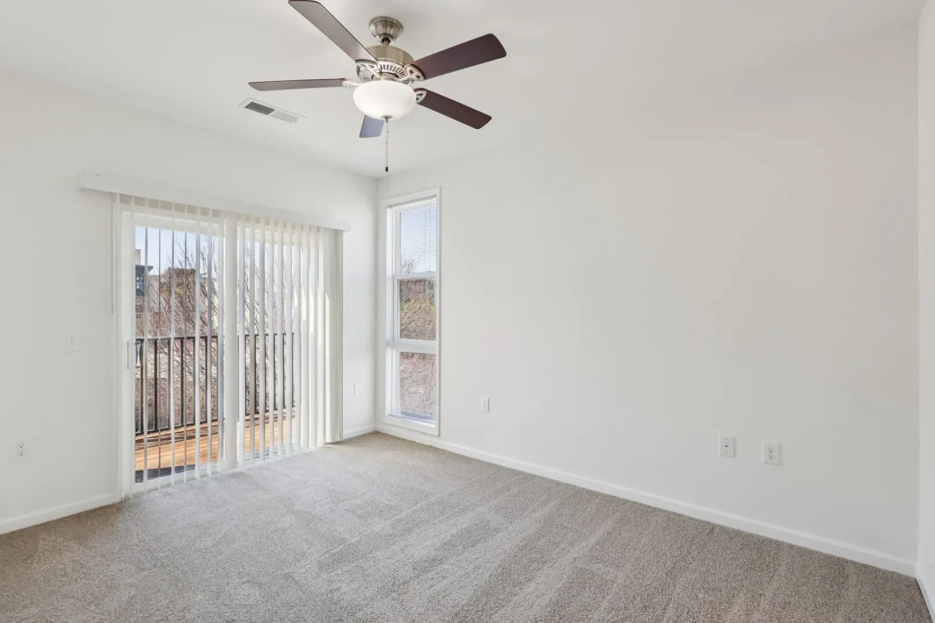 Empty apartment bedroom with neutral carpet, white walls, ceiling fan, and sliding glass doors to a balcony.