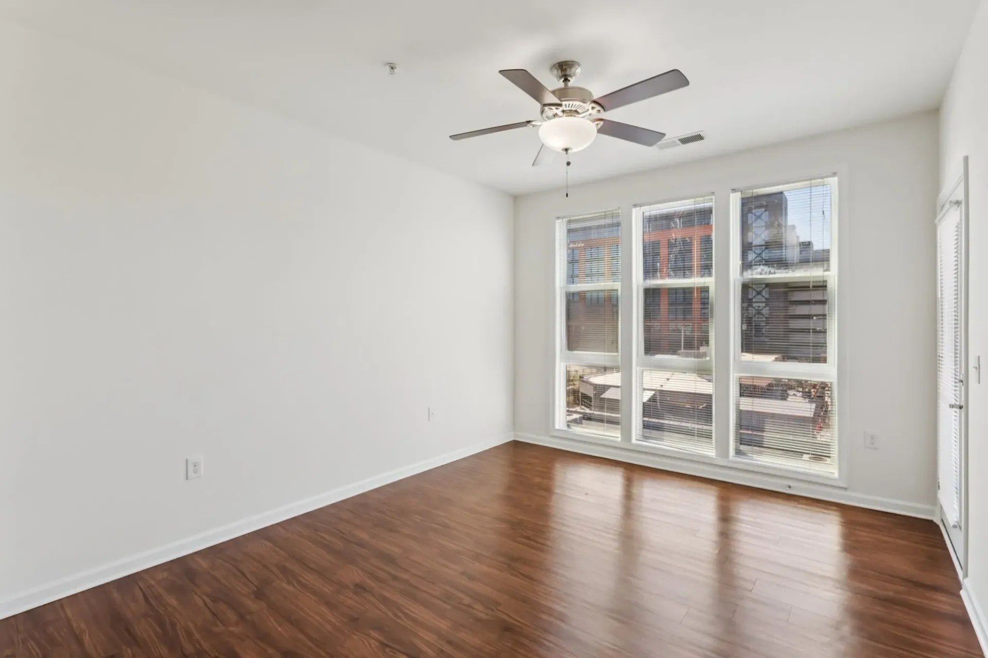 Empty living room with hardwood floors, a ceiling fan, and large windows overlooking the city.