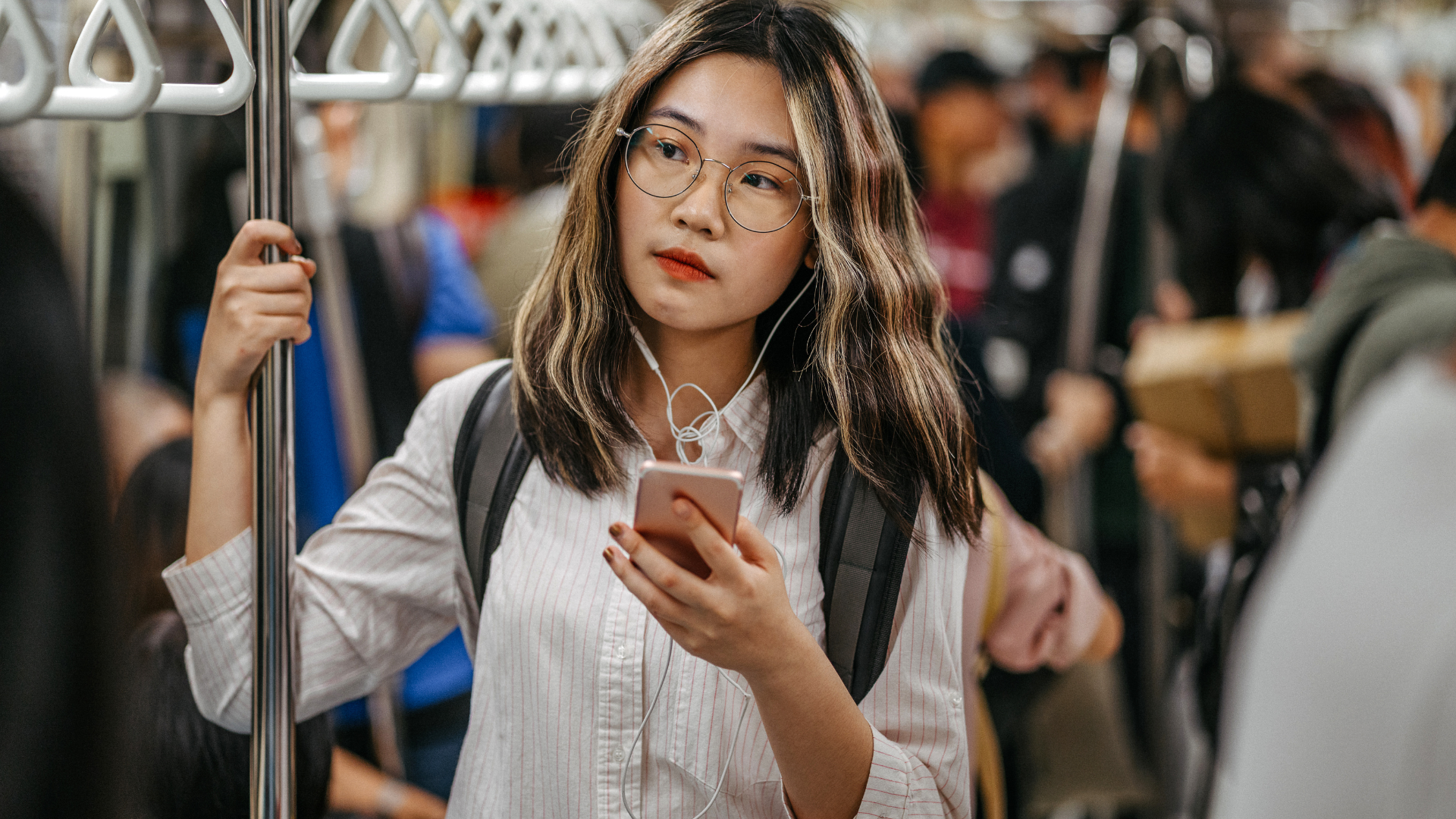 A person holding a phone and subway pole, wearing glasses and a light button-down shirt while standing in a crowded train.