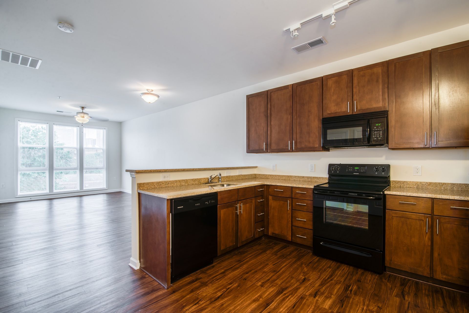 A kitchen with wood cabinets, black appliances, and granite countertops, opening into a living space with hardwood floors.