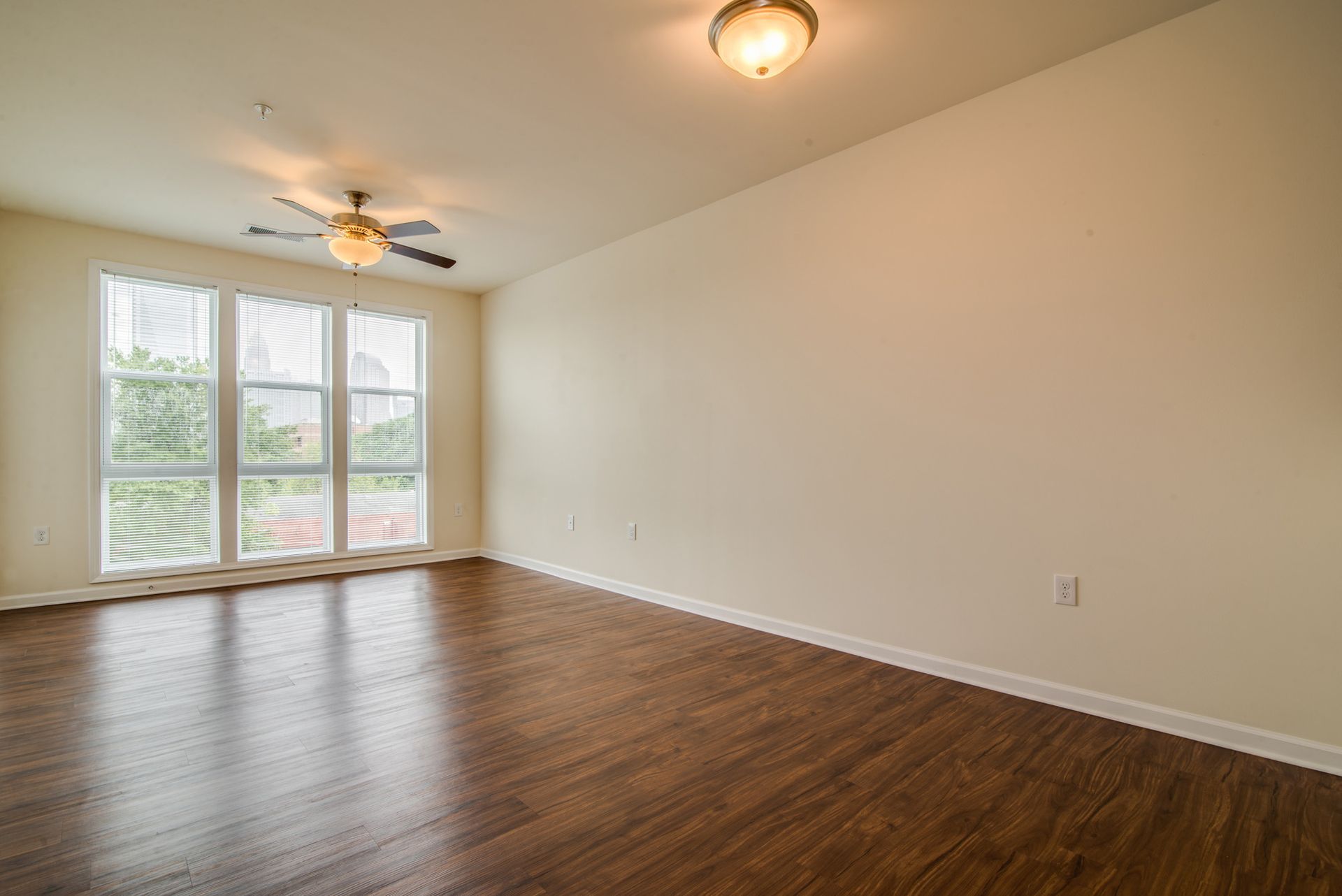 Empty room with wood floors, cream walls, large windows, and two overhead ceiling lights.
