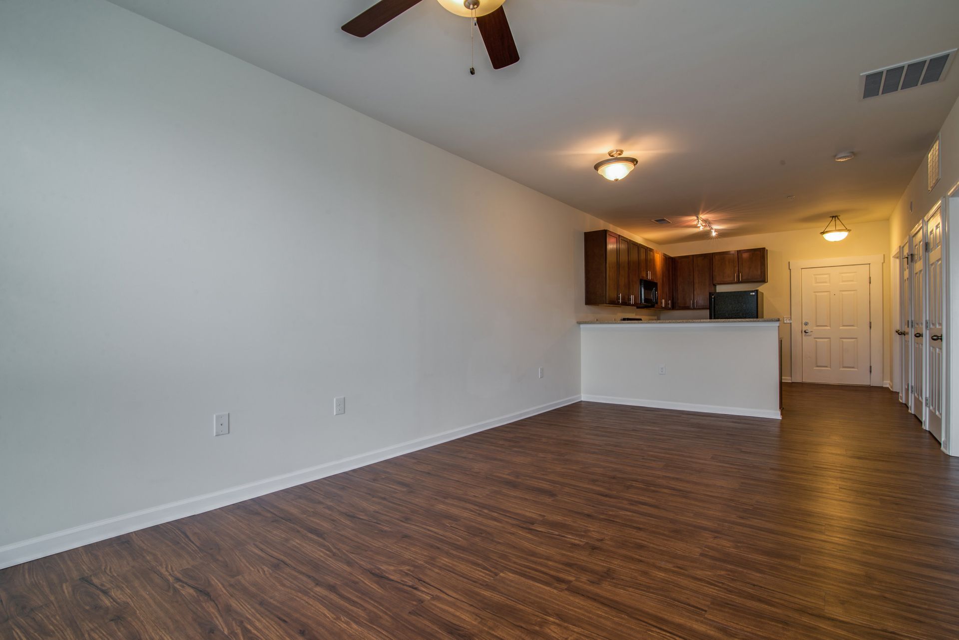 An empty living space featuring light walls, dark wood floors, a ceiling fan, and an open kitchen in the background.