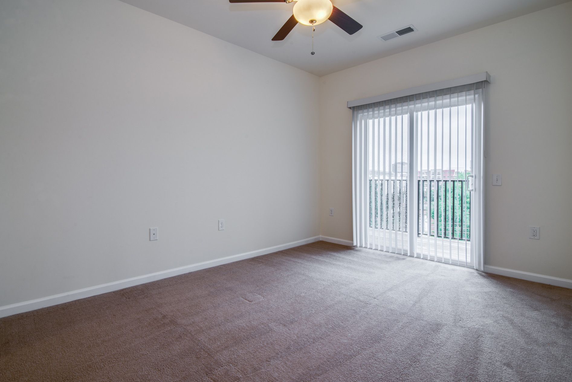 An empty room with beige carpet, light gray walls, a ceiling fan, and a sliding glass door leading to a balcony.