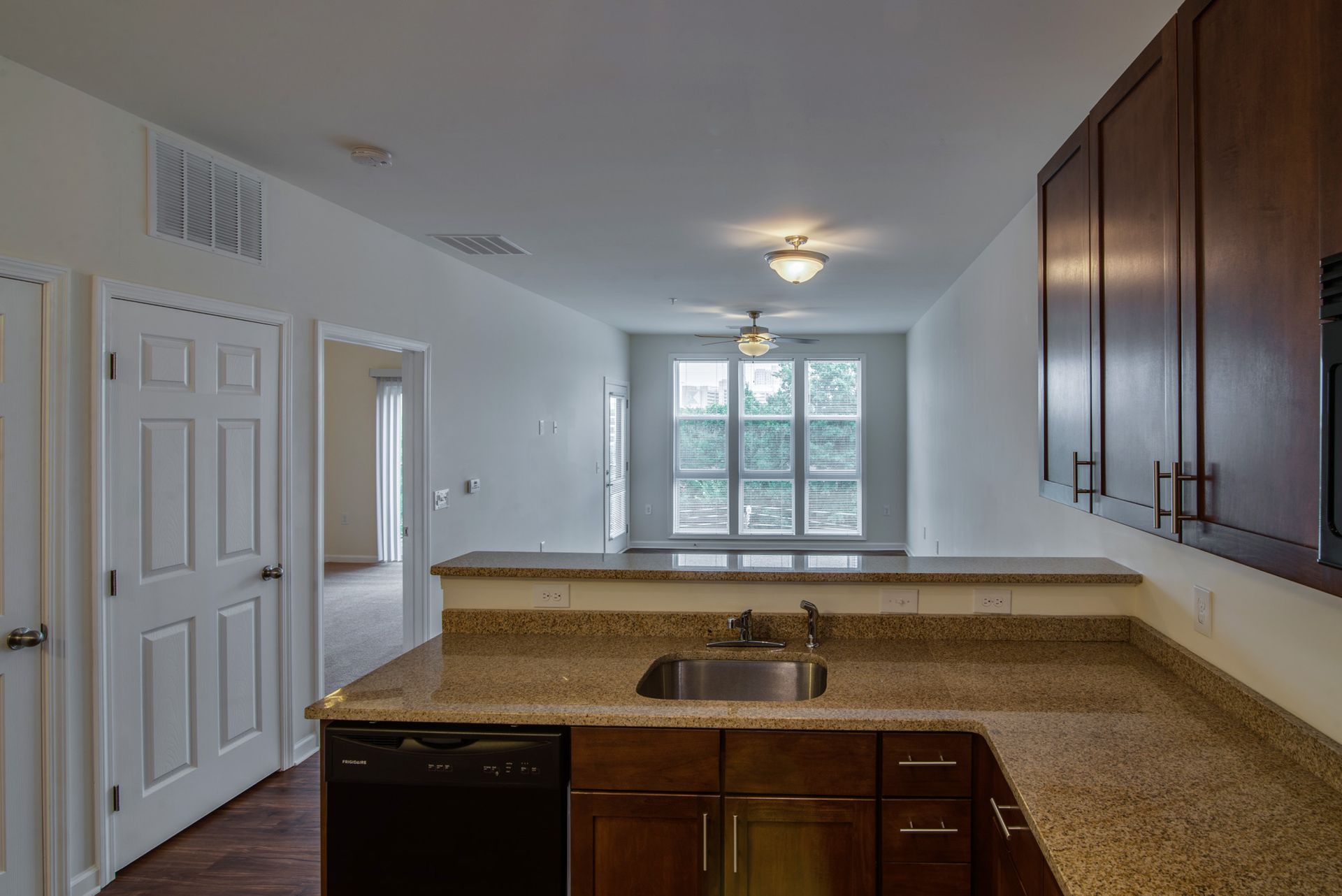 Kitchen sink area with granite countertops and dark wood cabinets, looking toward a sunlit living space and window.