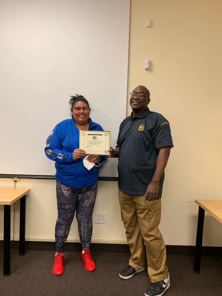 Woman in blue, holding a certificate, stands next to a man. They are in a classroom.