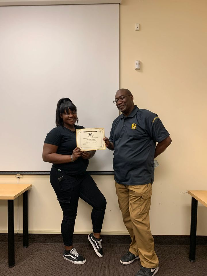 A person holds a certificate with another person in a room. Both smile. They stand by a whiteboard and tables.