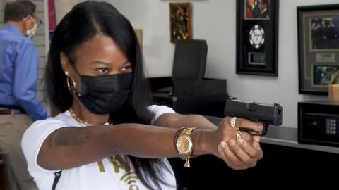 Woman aiming a handgun, wearing a black mask and gold jewelry, indoors.