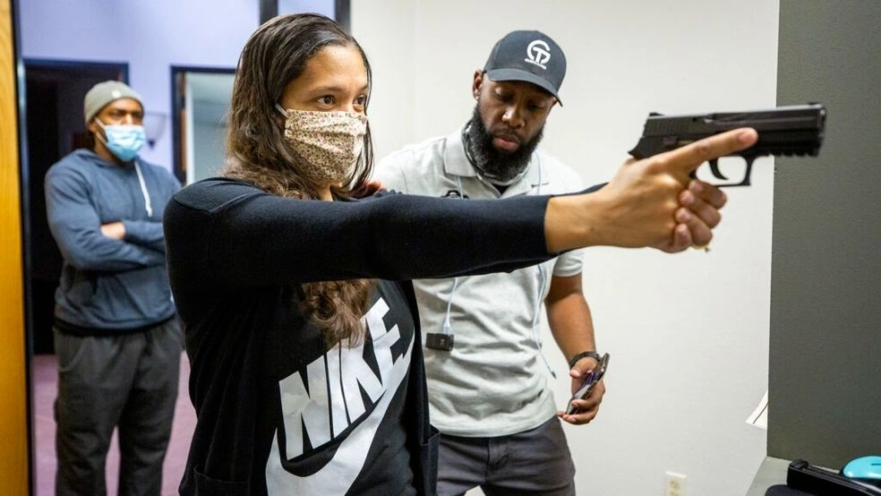 Woman aims a handgun during a training session. Instructor observes as another person waits in the background.