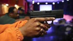 Person holding a black handgun, pointing forward, with others in the blurry background.