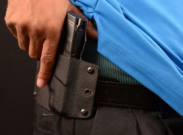 Person's hand drawing a pistol from a black holster clipped to a belt. Blue shirt, black background.
