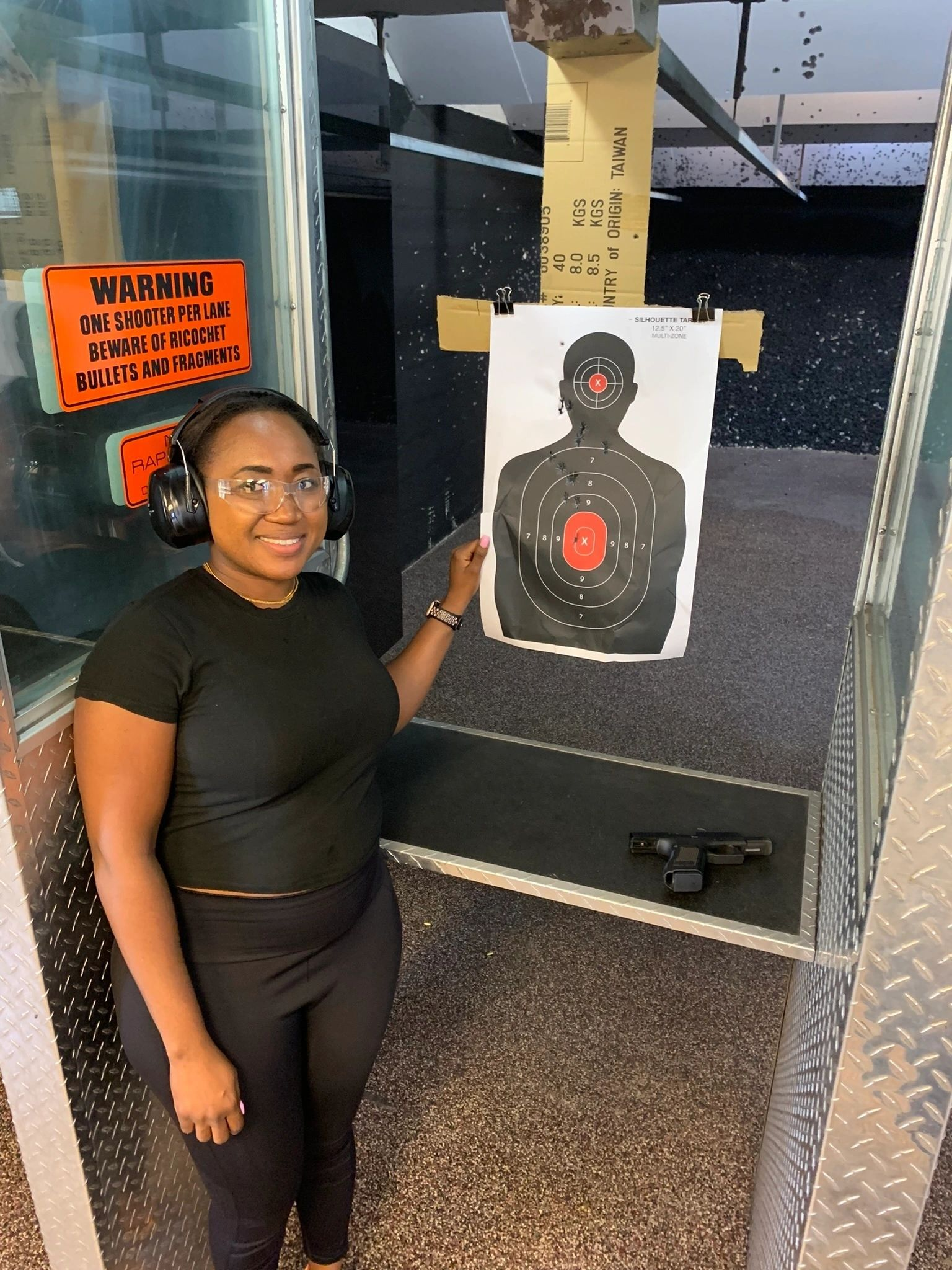 Woman in earmuffs at shooting range, holding target with bullet holes. Pistol on table.
