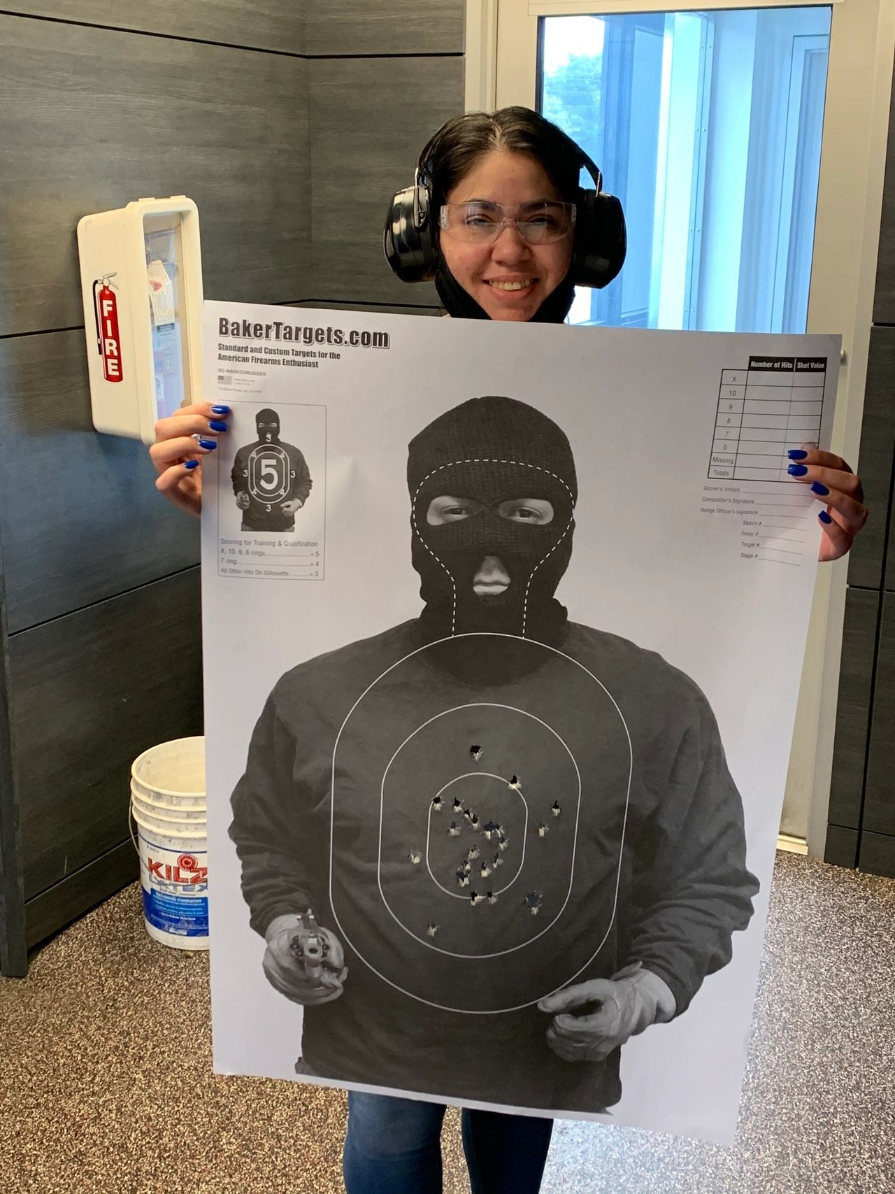 Woman holding a target with bullet holes in a shooting range, wearing ear protection and smiling.