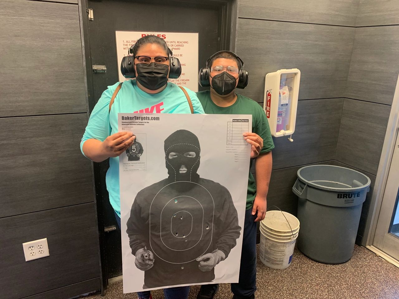 Two people at a shooting range hold a target with bullet holes. They wear ear protection and masks.