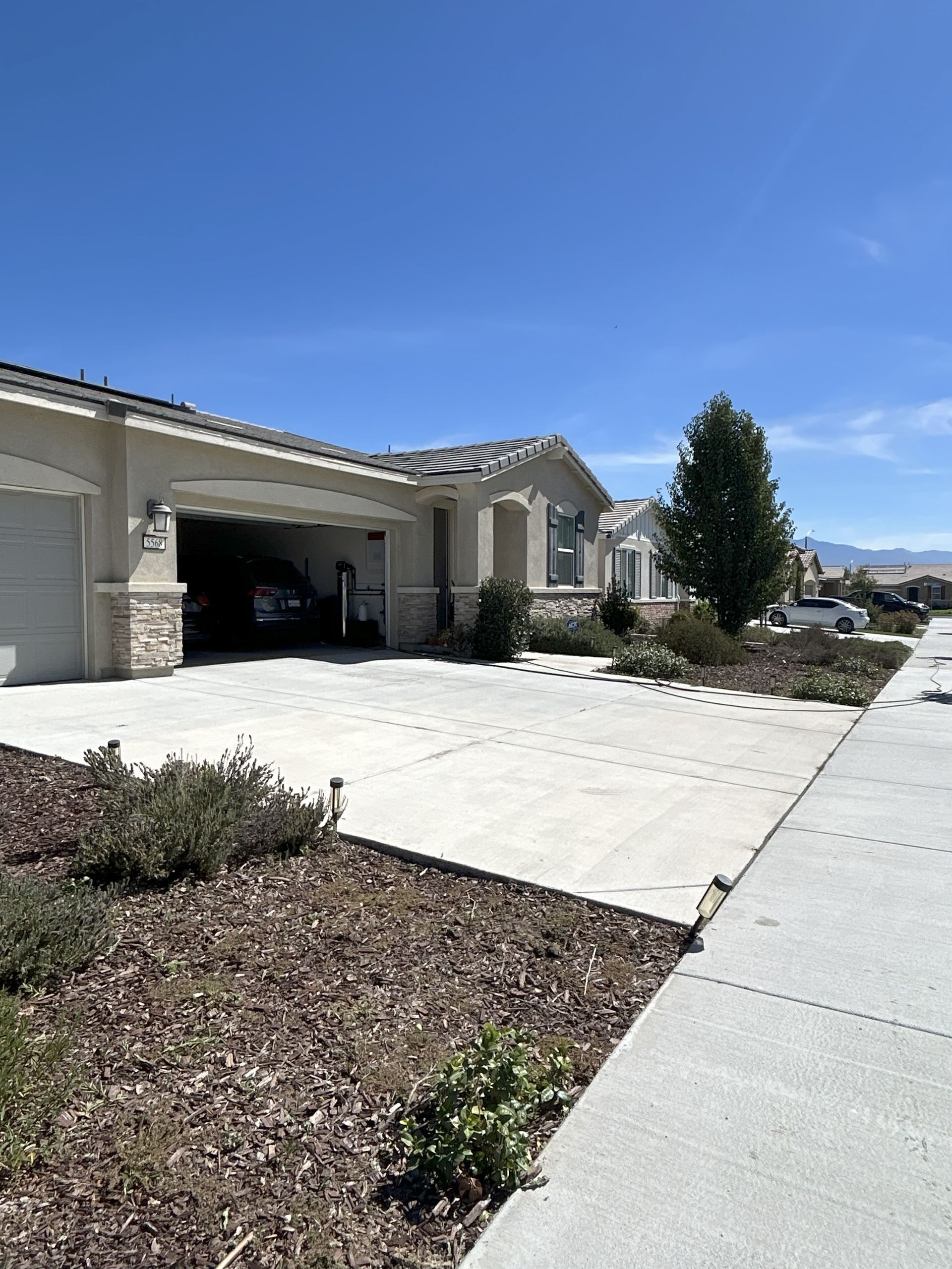 Beige house with open garage, driveway, sidewalk, and small front yard landscaping under a blue sky.