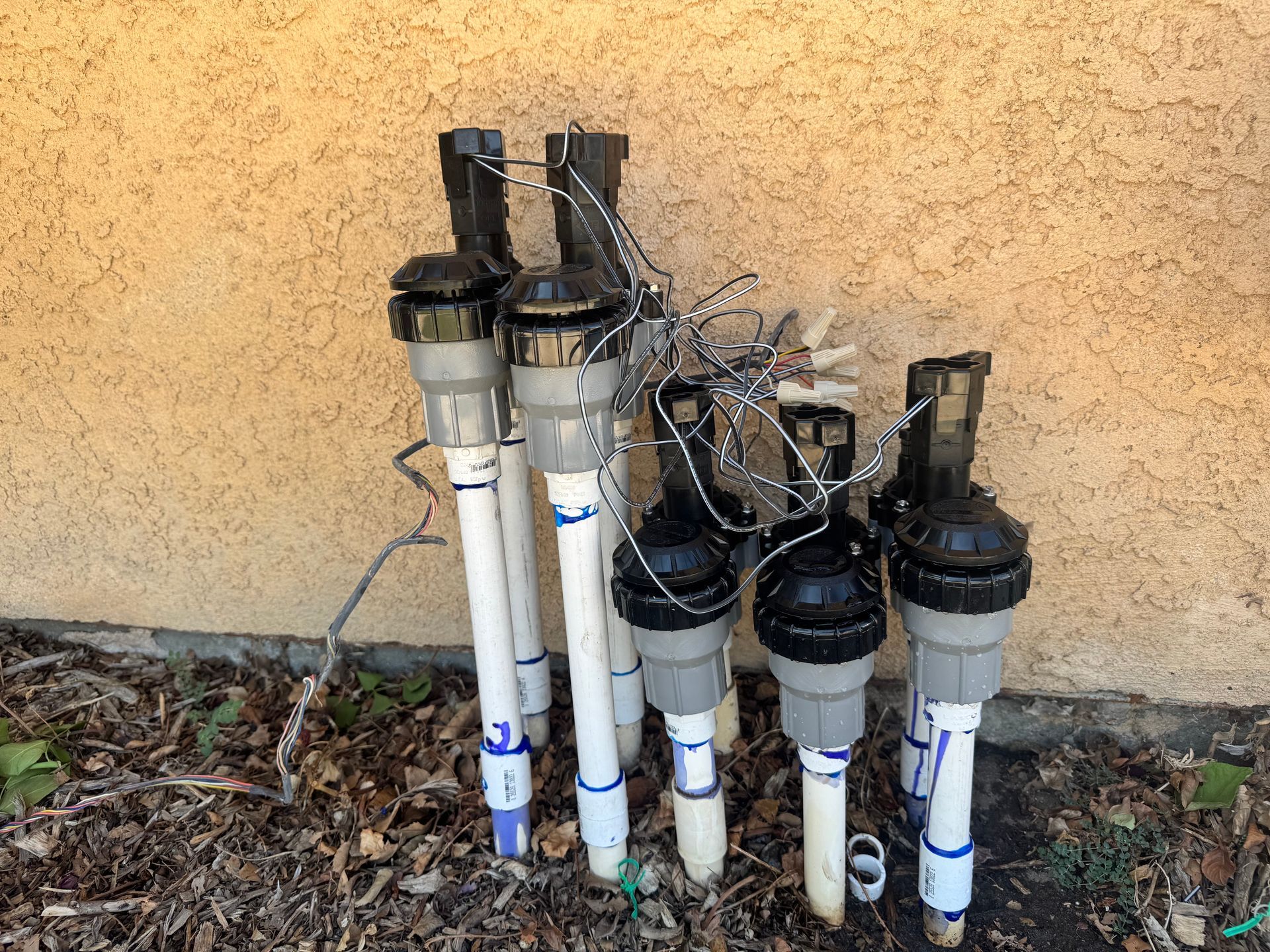 Sprinkler system valves against a beige stucco wall, with tangled wires and plastic pipes.