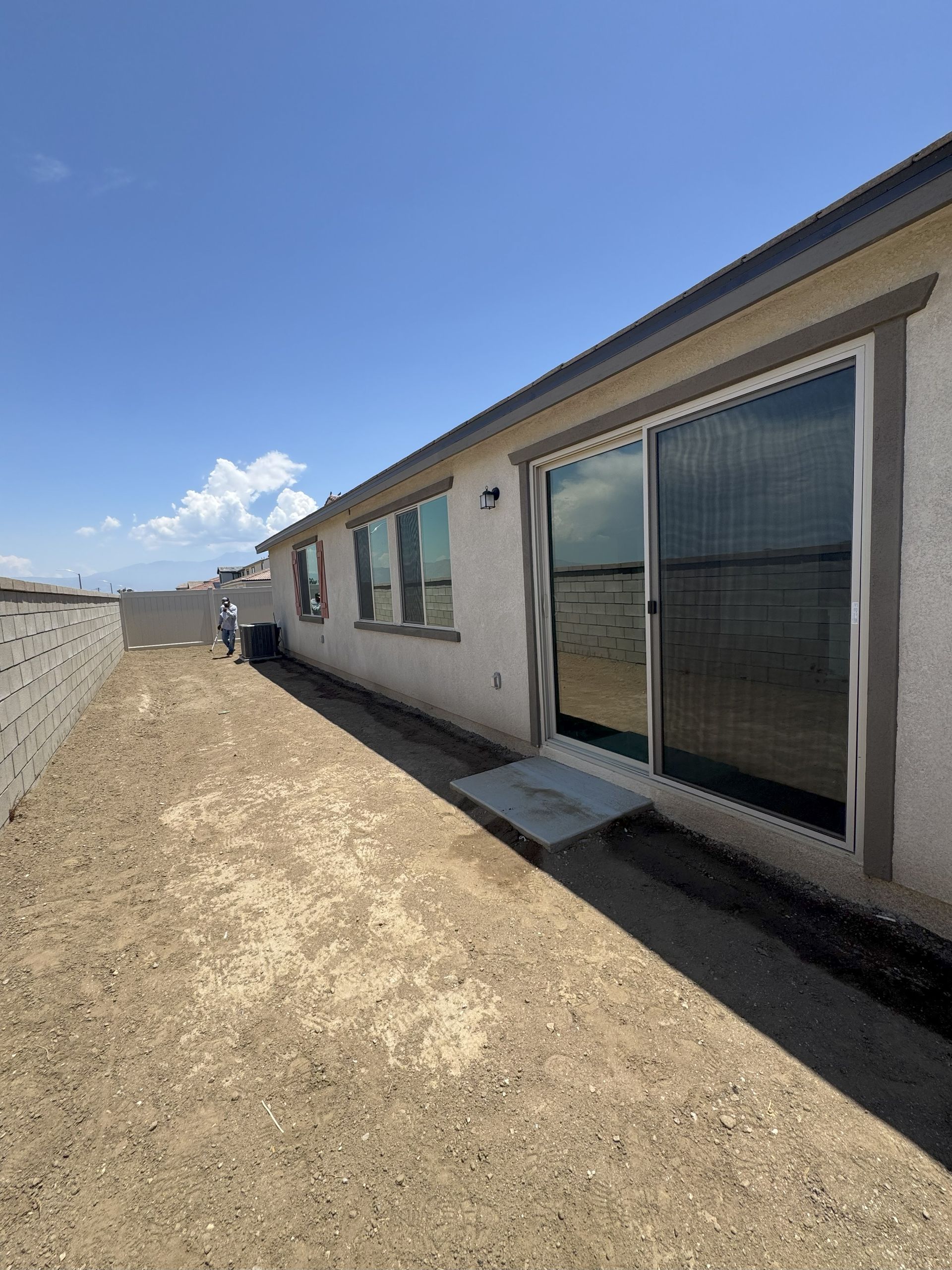 Side view of a beige house with a sliding glass door, ramp, and bare dirt yard on a sunny day.