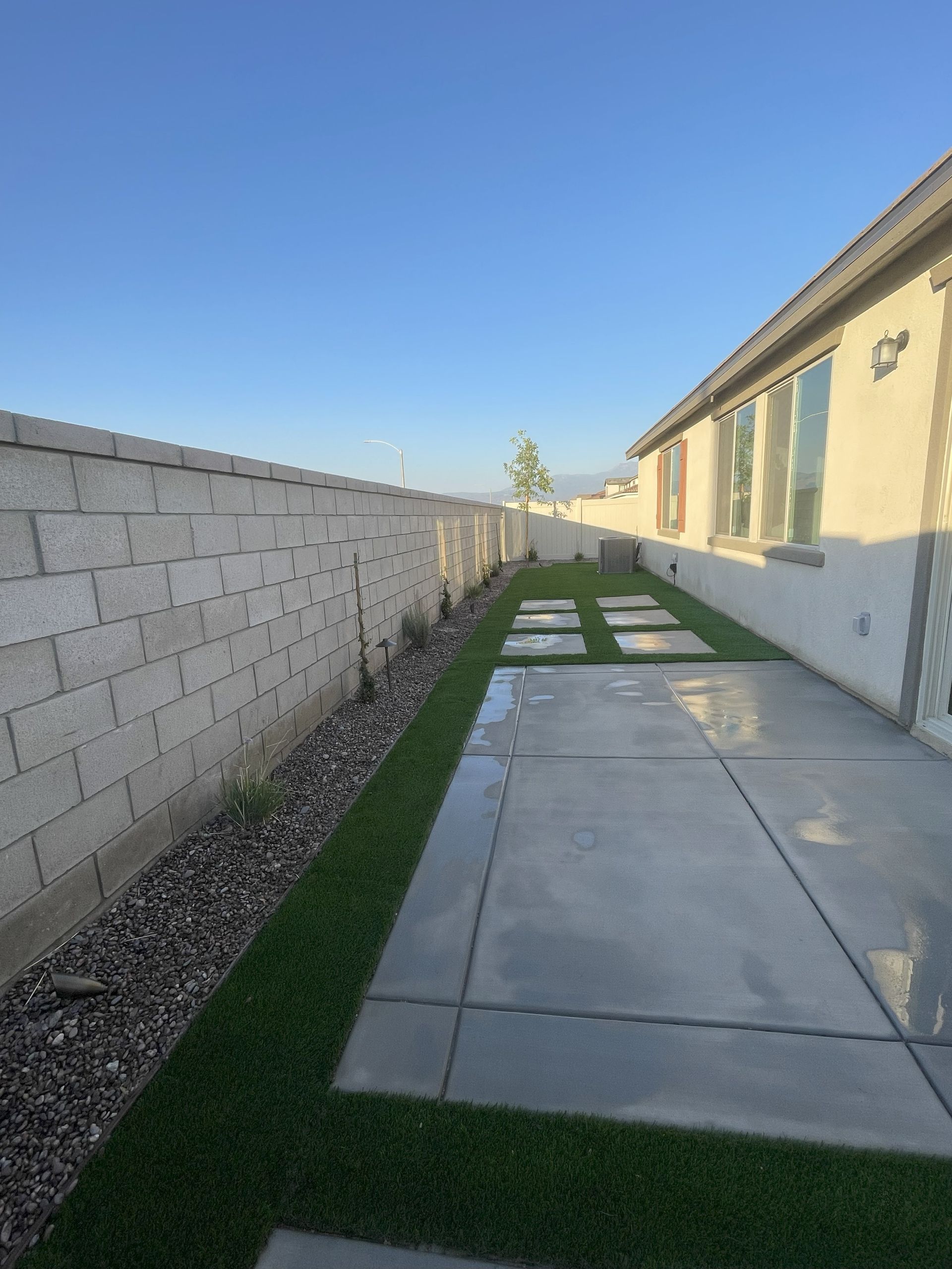 Backyard with concrete patio, grass, and block wall under a blue sky.