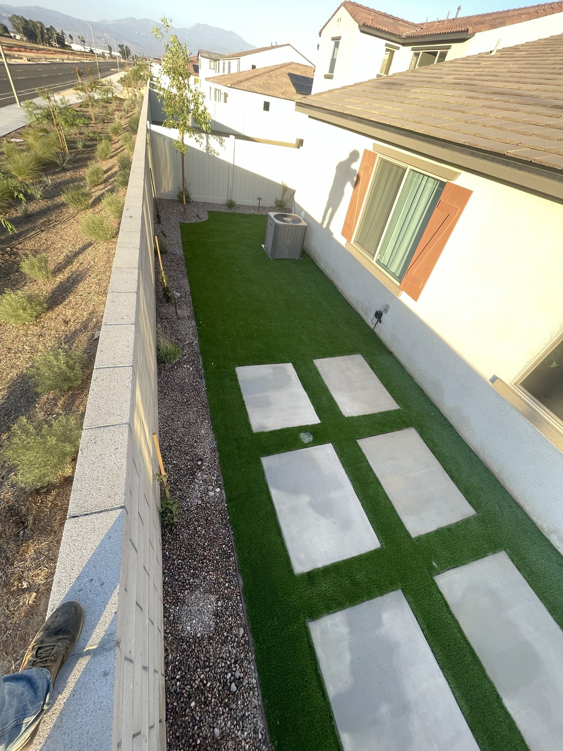 Narrow backyard with artificial turf and stone pavers bordered by a concrete block wall, in front of a house.