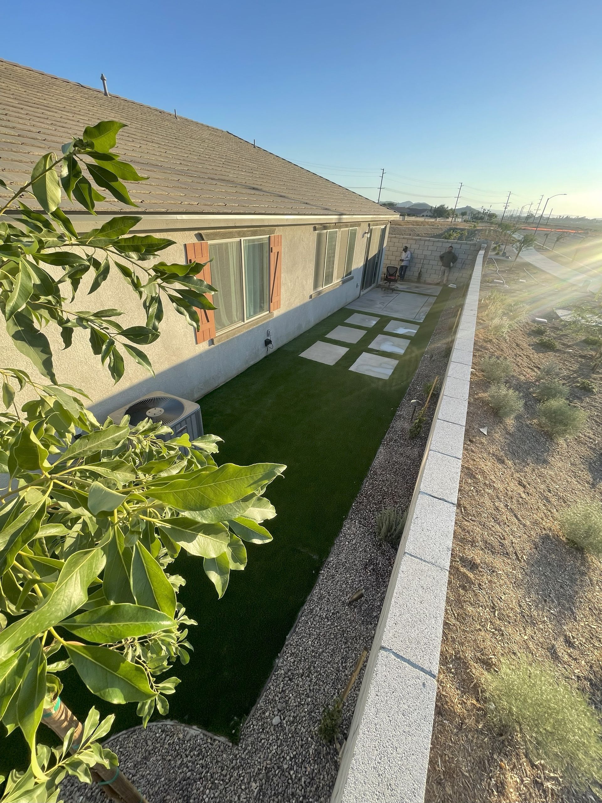 Side view of a house with a grassy lawn, concrete pavers, and a stone wall under a sunny sky.