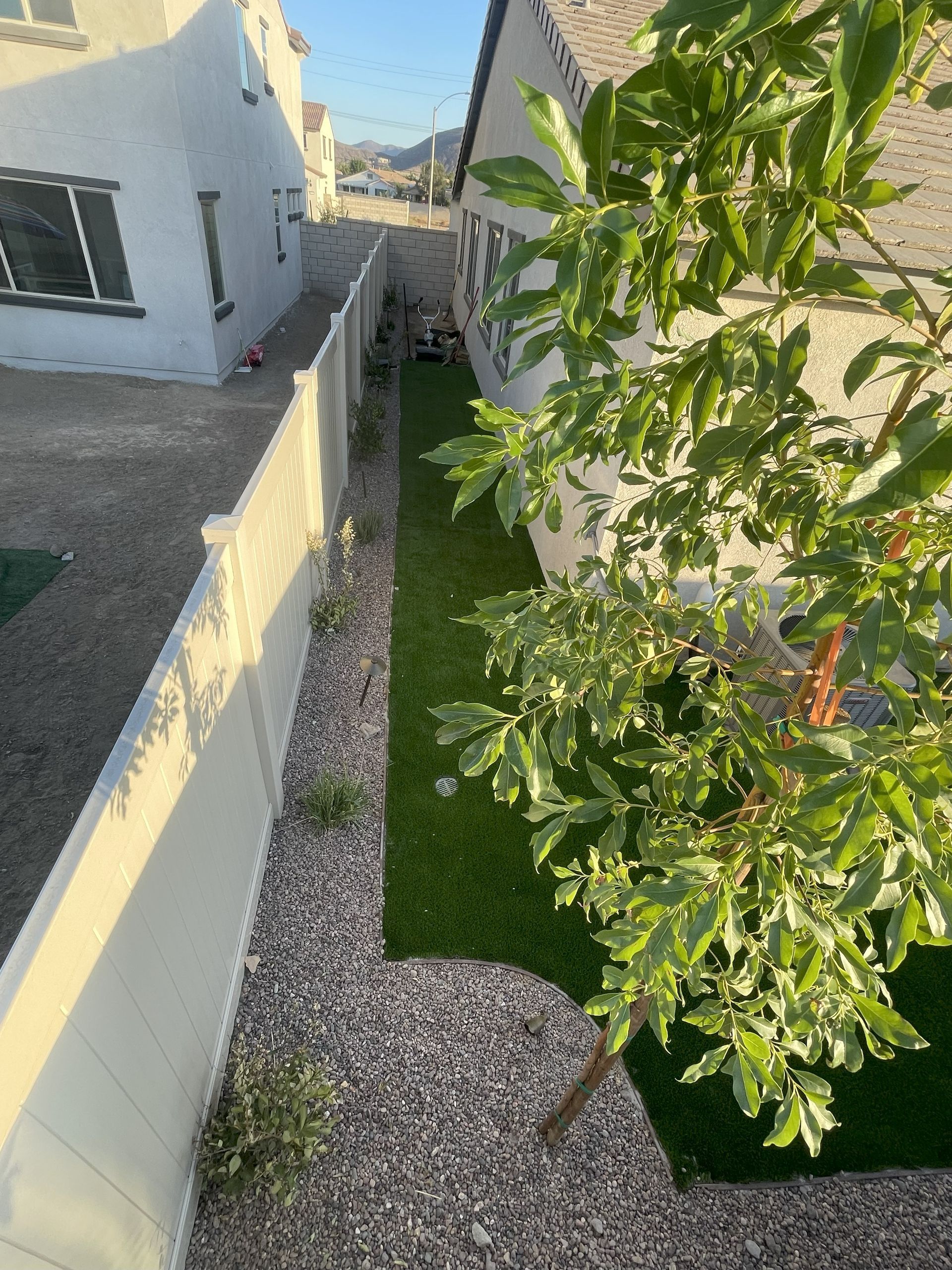 Narrow backyard with gravel, fake turf, white fence, and a tree.