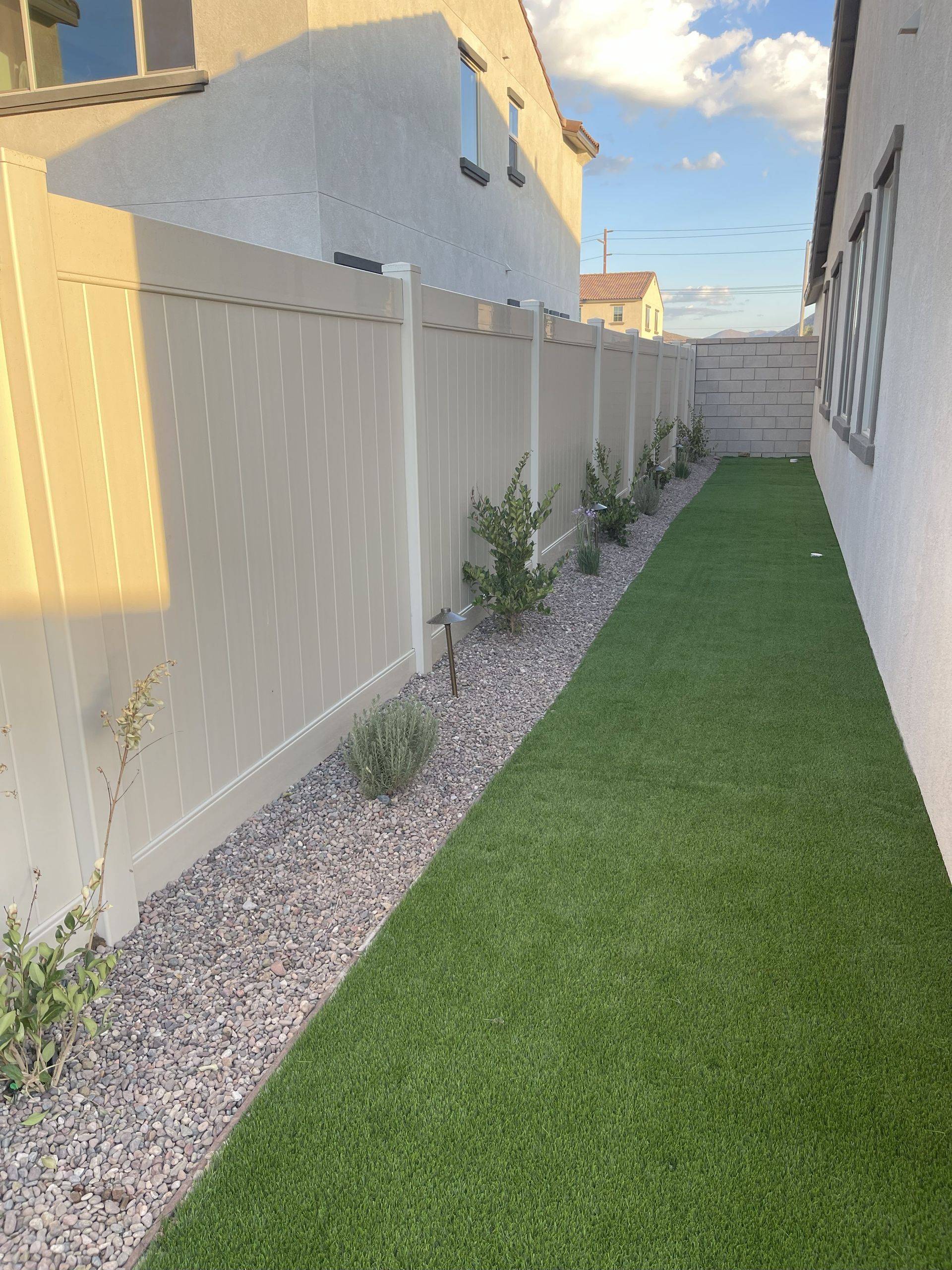 Narrow backyard with beige fence, green turf, gravel border, and small plants.
