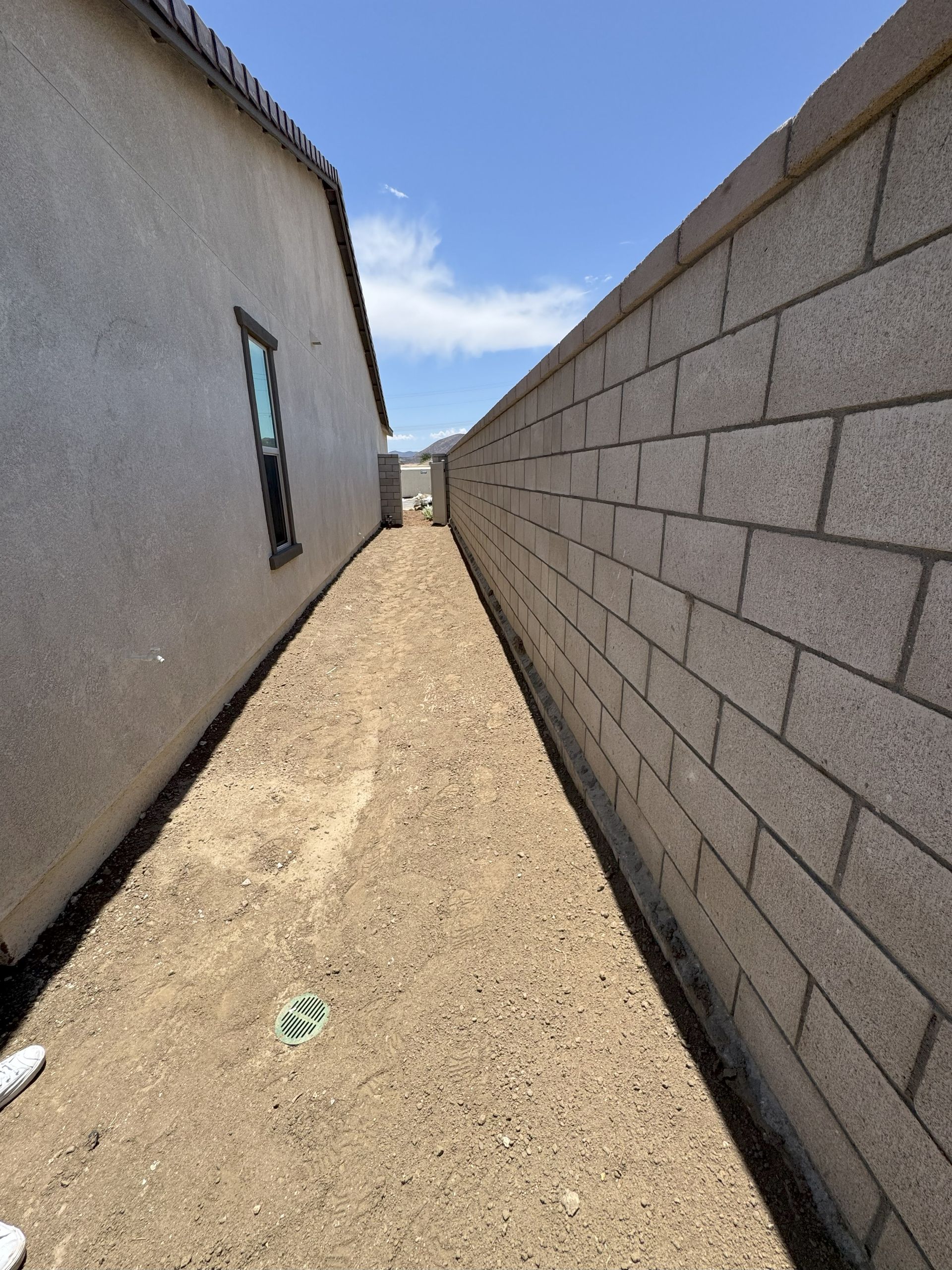 Narrow dirt path between a stucco house and a block wall under a blue sky.