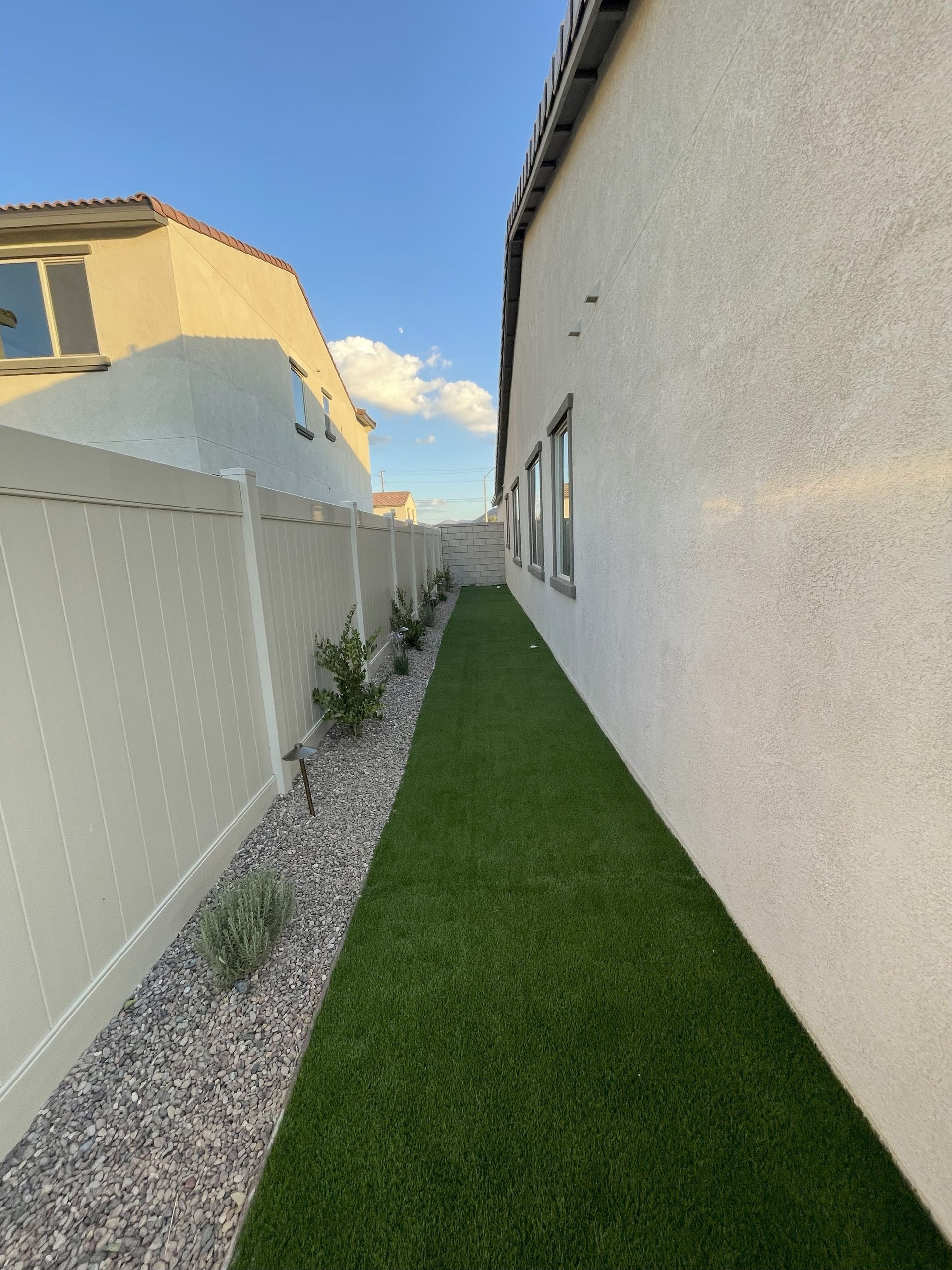 Narrow backyard with green turf, white fence, beige house, and a blue sky.