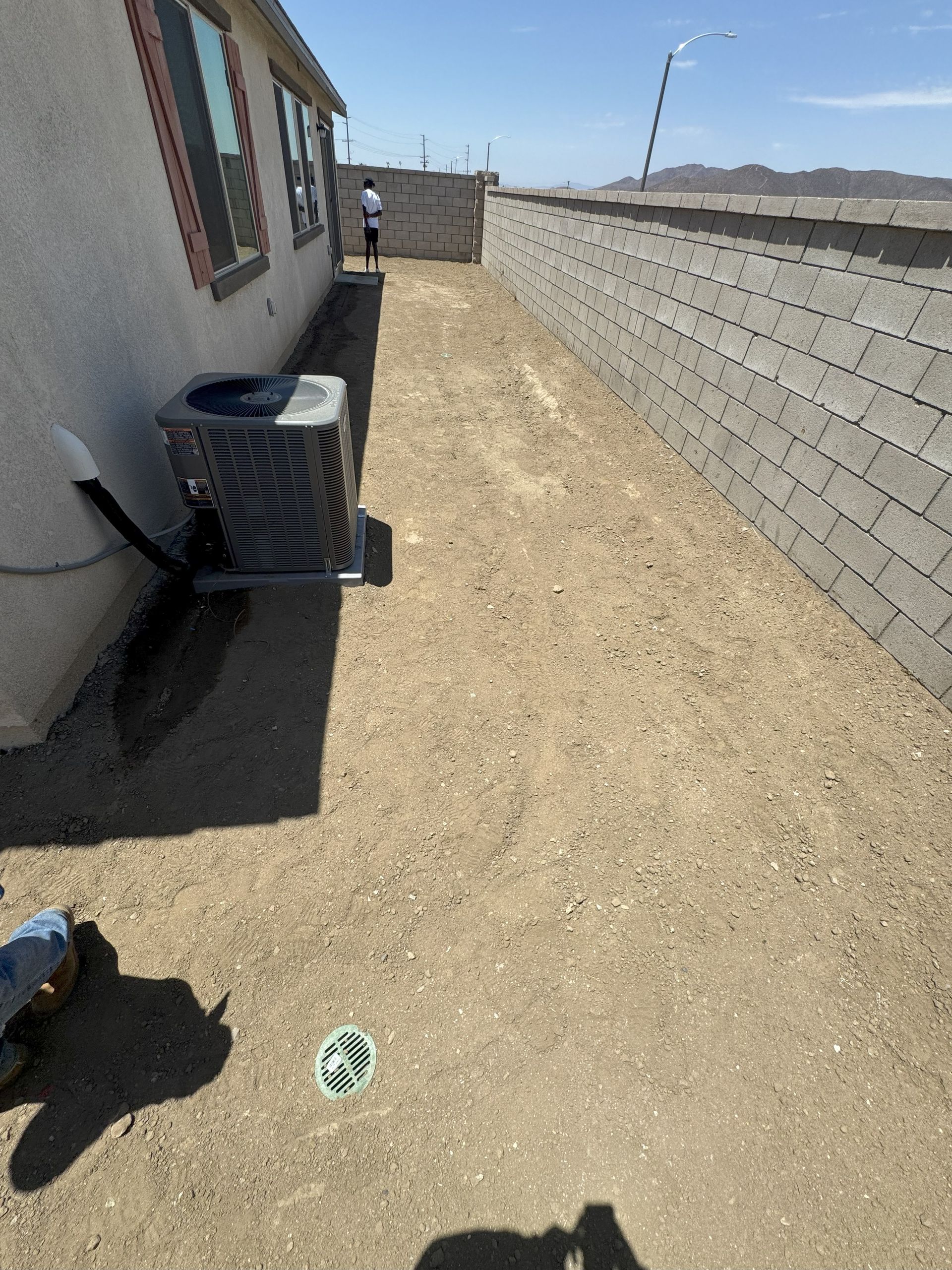 A side yard with an AC unit, a wall, and a person in the distance, all on dirt.