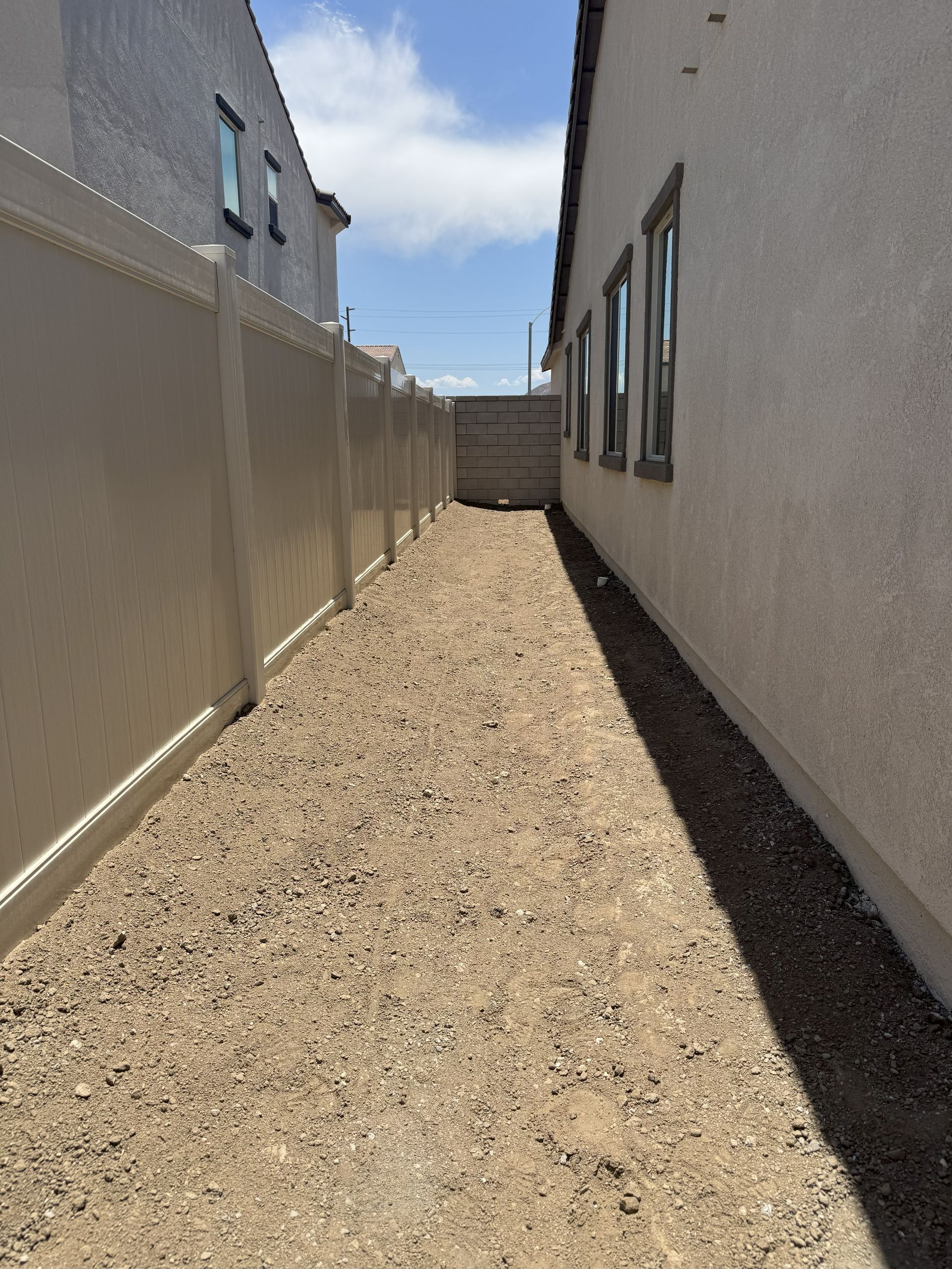 Narrow gravel pathway between a beige fence and a stucco house, under a bright sky.