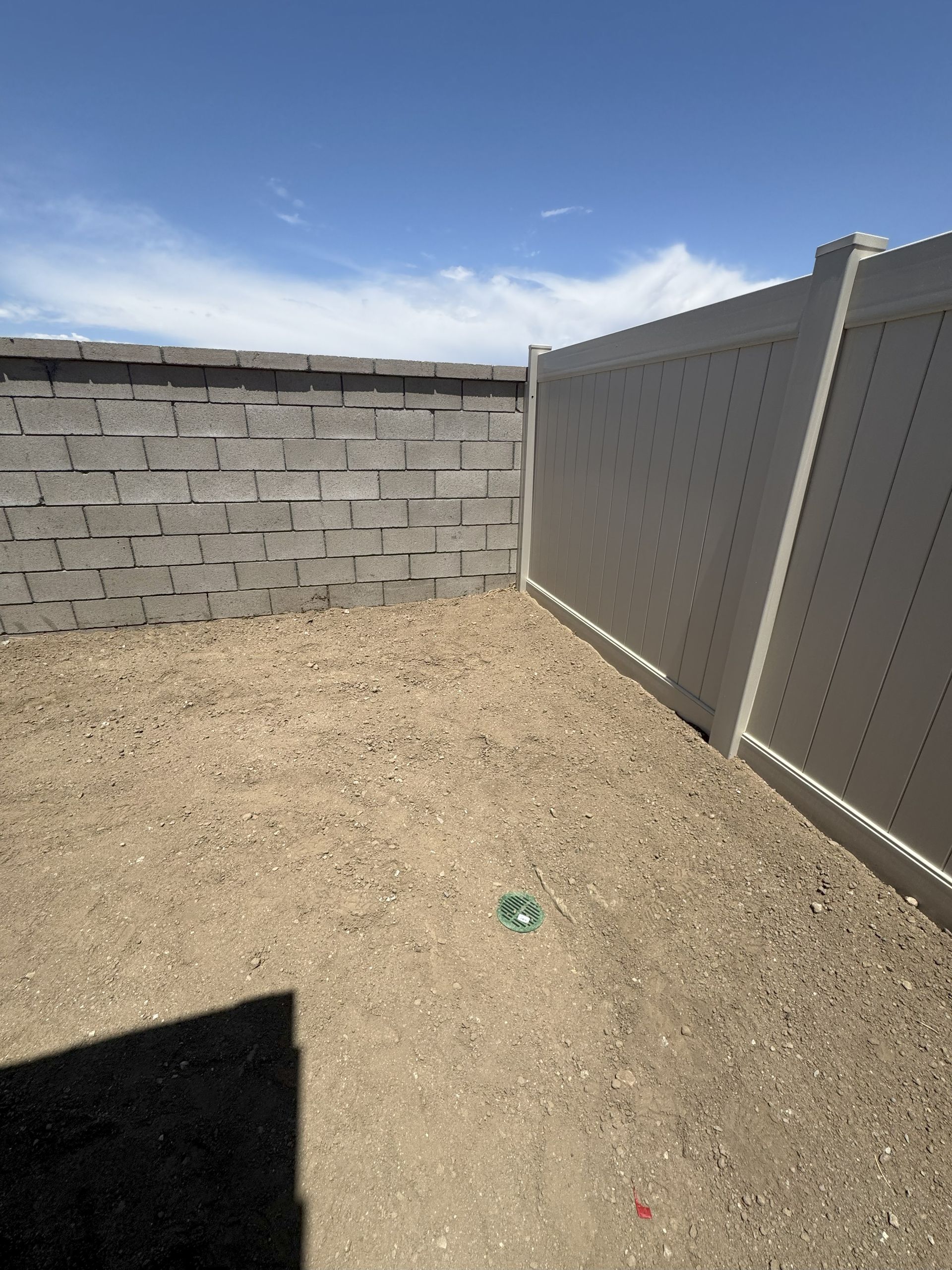 A gravel backyard enclosed by a grey brick wall and a white vinyl fence under a blue sky.