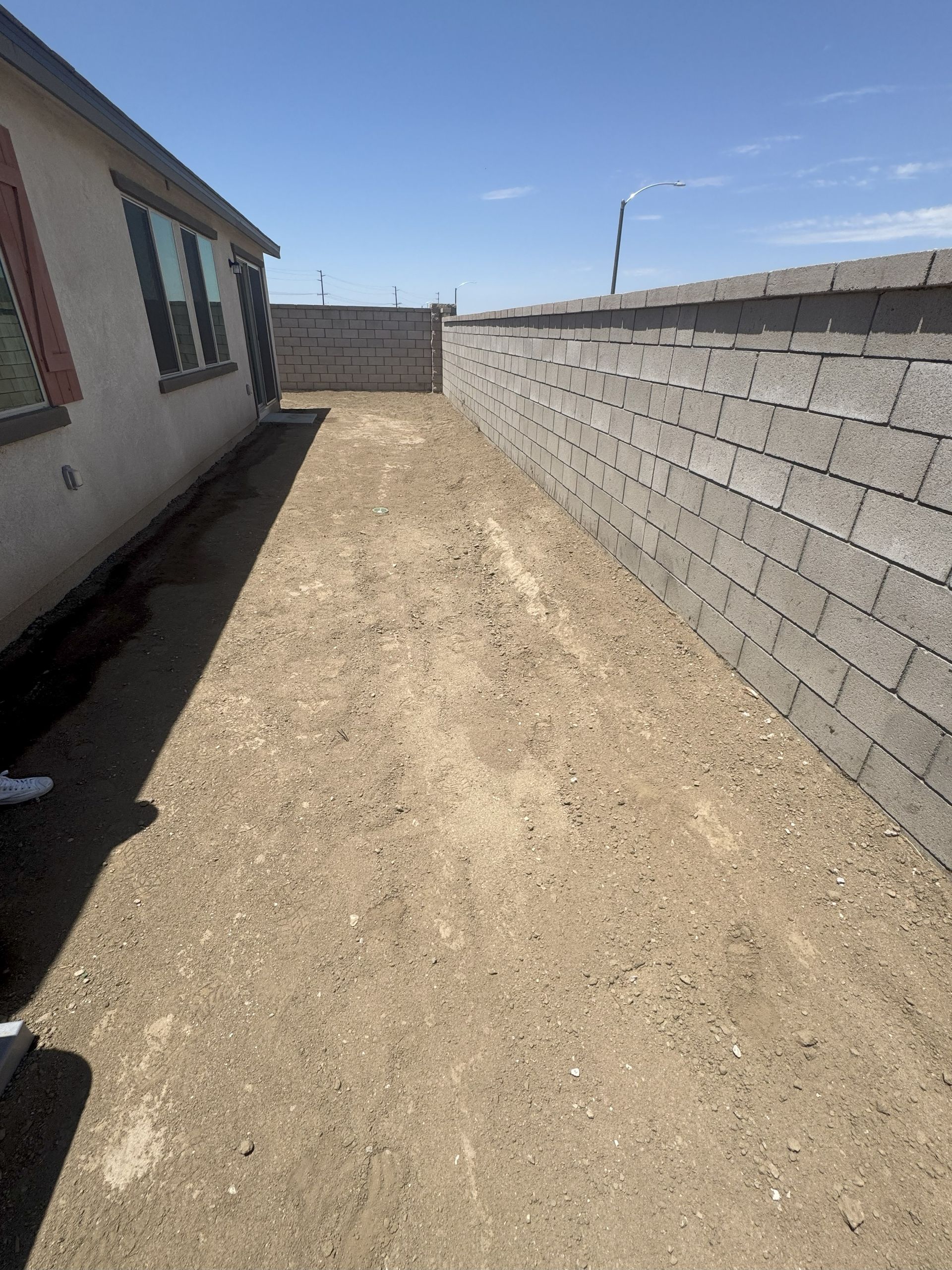 Backyard with dirt path between house and block wall, under a clear blue sky.