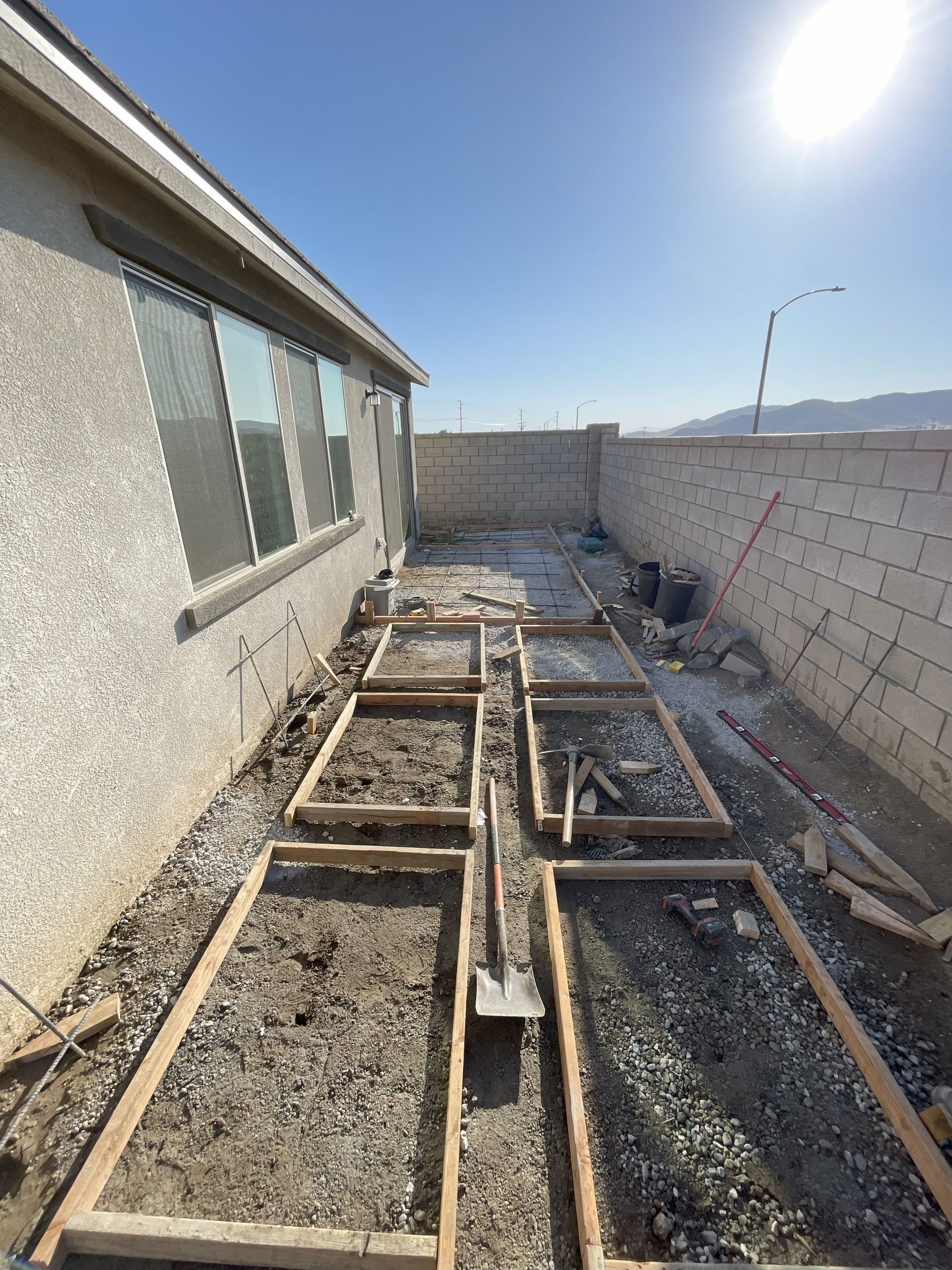 A construction site with wooden frames on gravel ground. A house is on the left, and a wall is on the right.